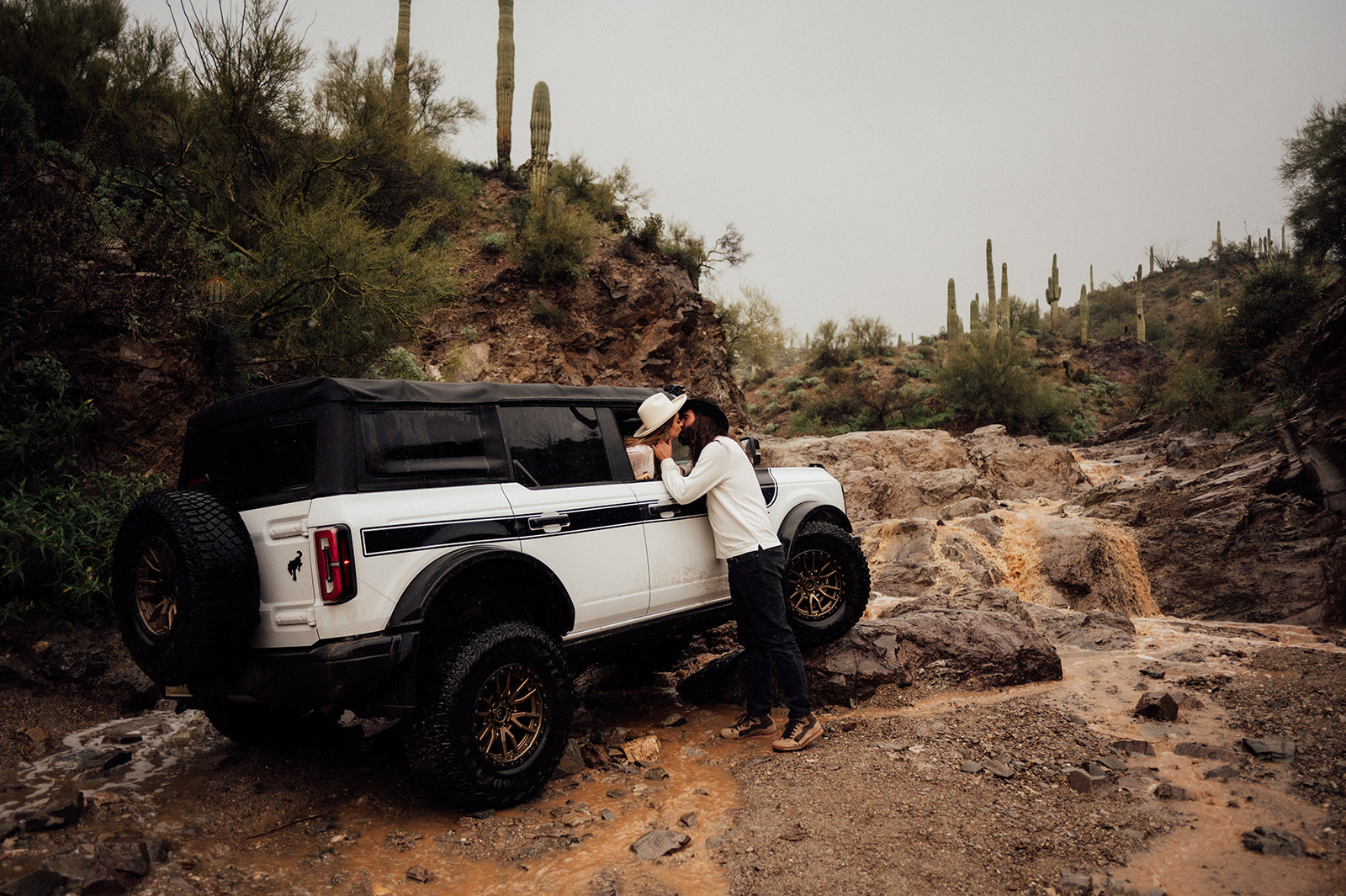 Groom leaning into window of Ford Bronco kissing his bride in front of a waterfall in an Arizona canyon