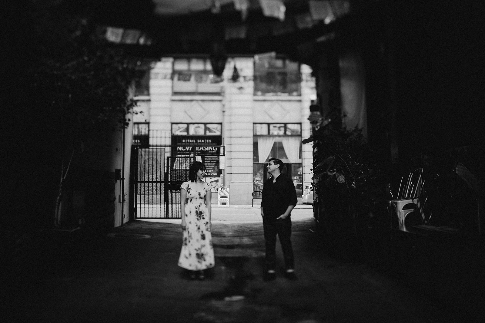 black and white photo of couple standing apart in an alley with natural light on them
