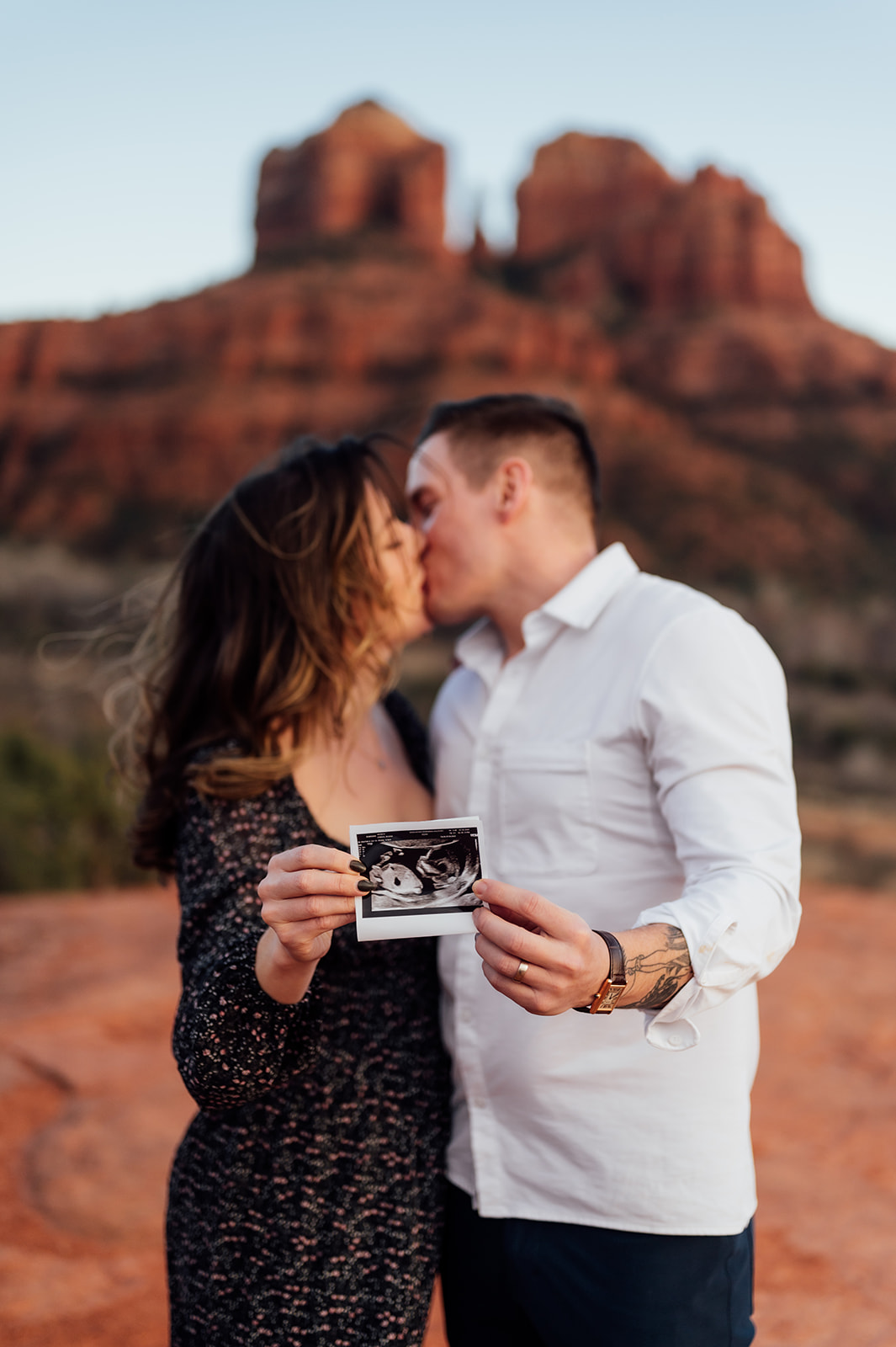 couple holding up a sonogram photo of their baby girl while kissing in front of Cathedral Rock in Sedona