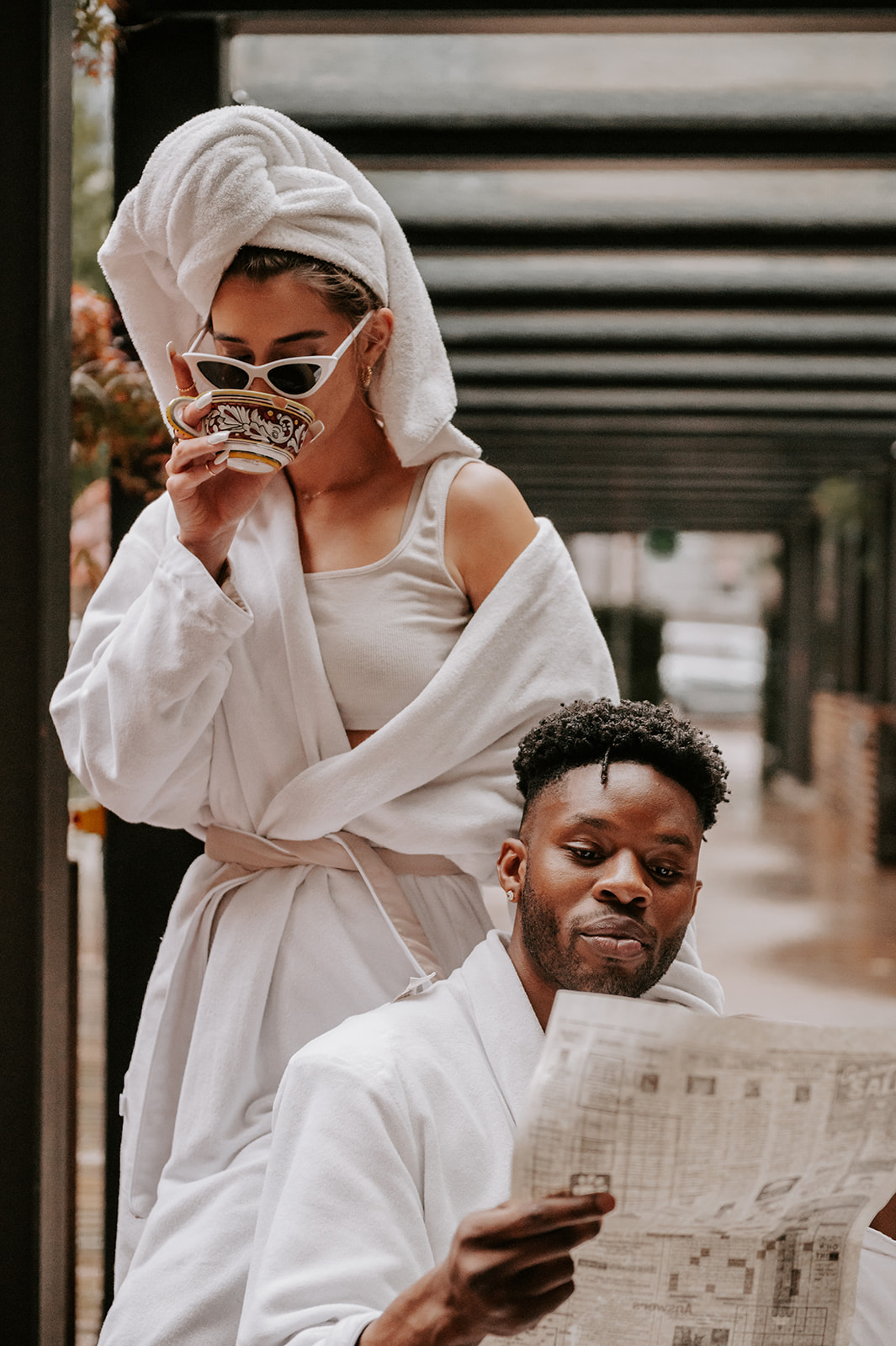 Couple embracing and reading the paper together. Wearing robes and she has a towel on her head and white sunglasses
