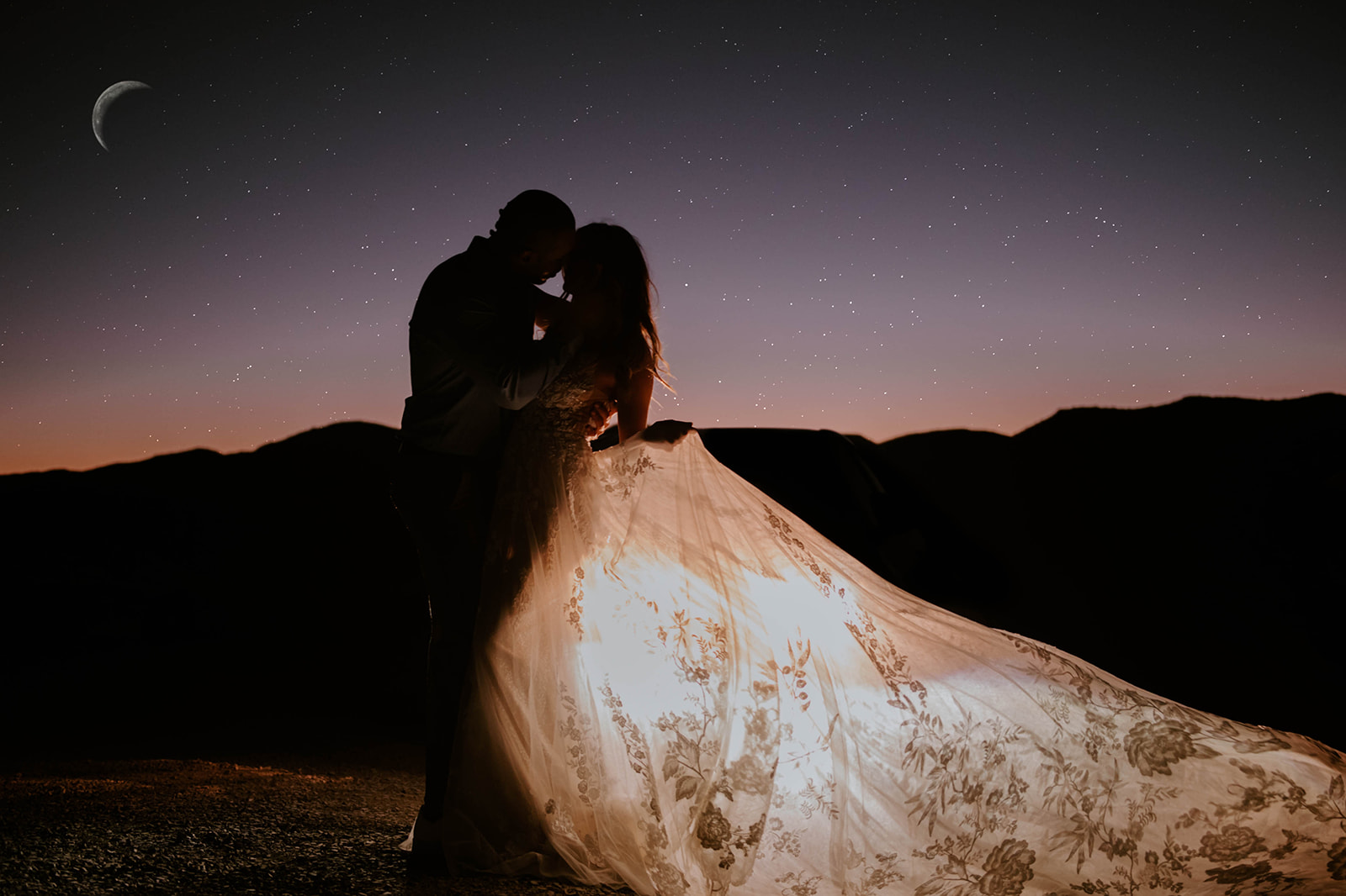 A bride and groom standing face to face silhouetted under the moon and stars night sky with her dress lit up