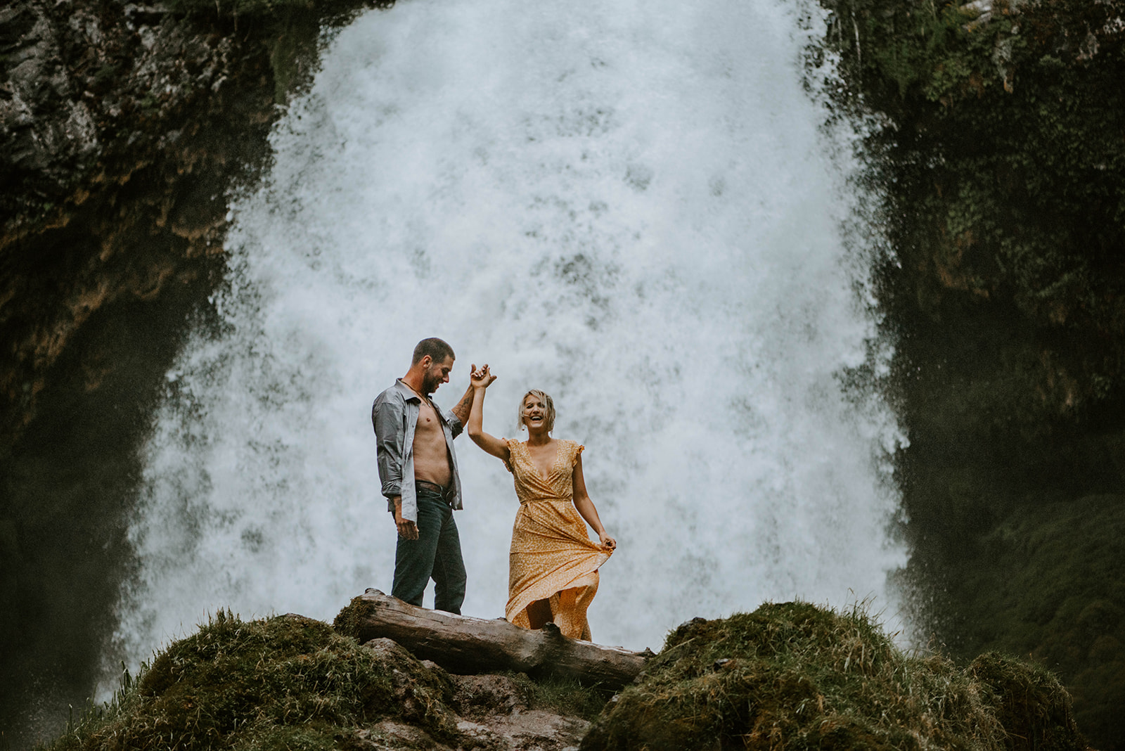 A couple laughing together in front of Sahalie Falls during their engagement photos. She is wearing a yellow dress 