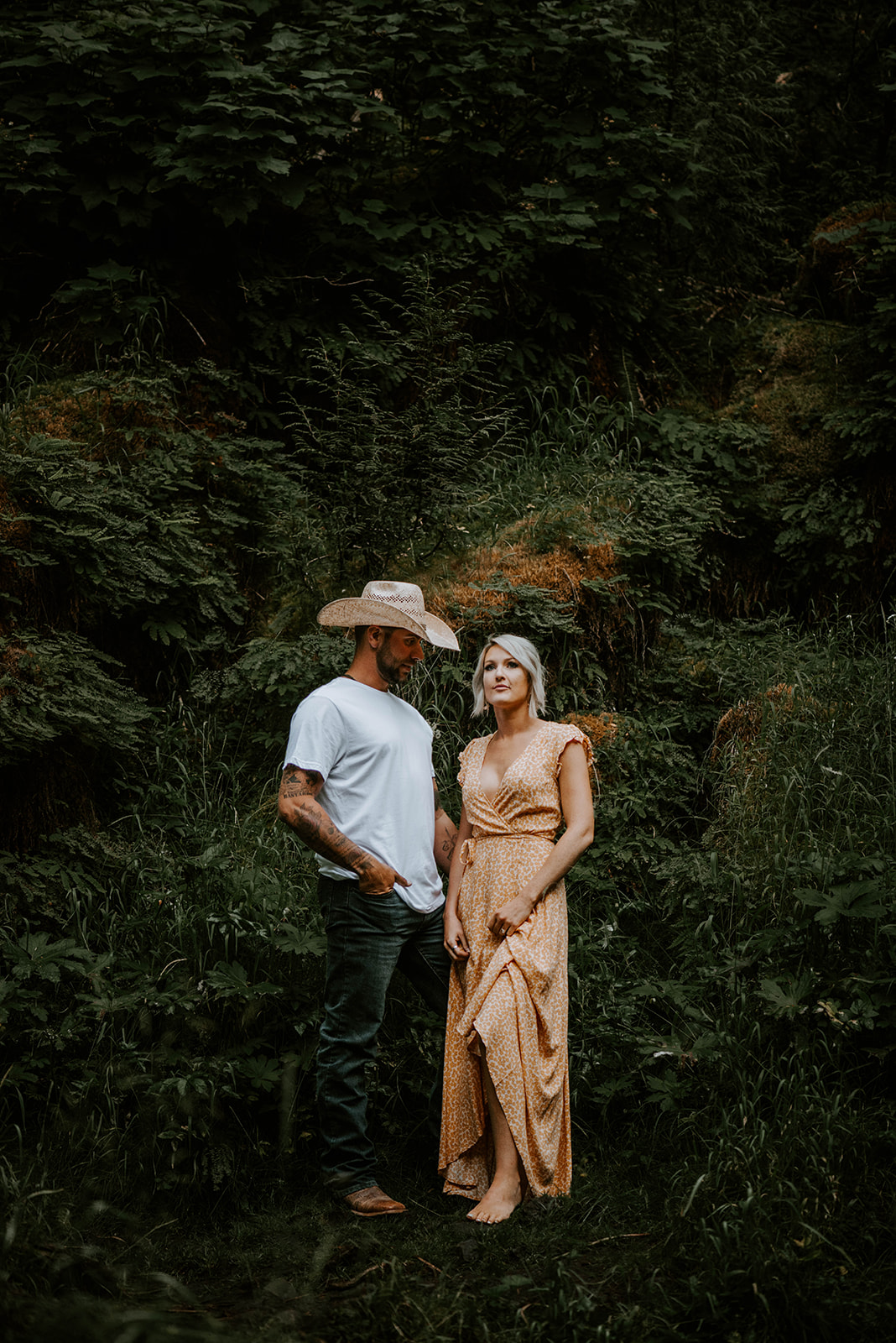 A couple posing against a green wall for their engagement photos. The guy is wearing a cowboy hat.
