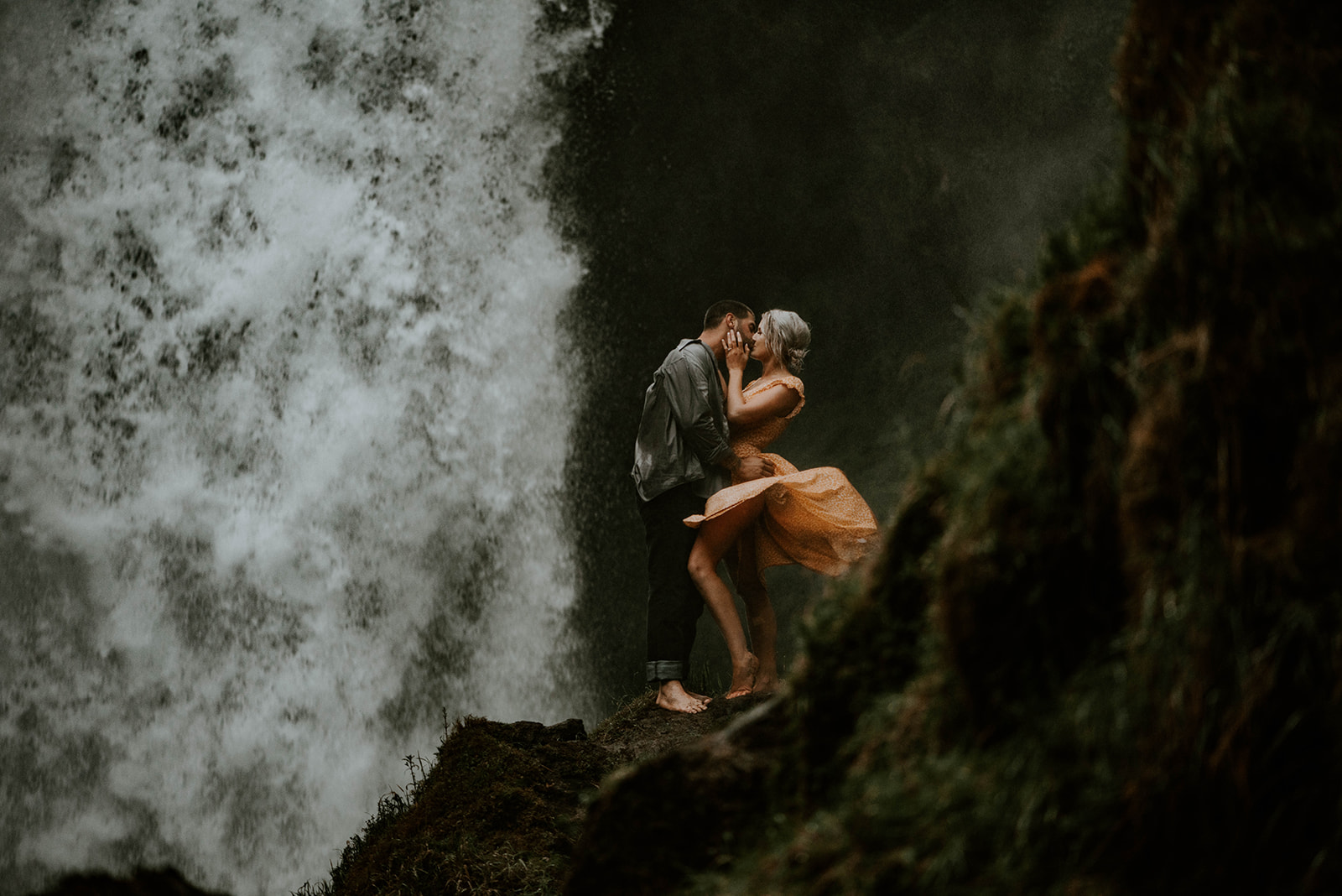 A woman in a yellow dress and a man in a grey shirt kissing under a waterfall while her dress blows