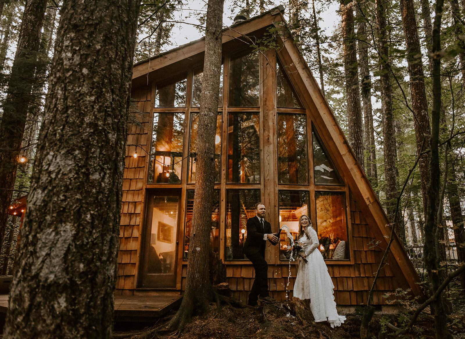 A couple who eloped popping a bottle of champagne in front of an A-Frame cabin after saying their vows