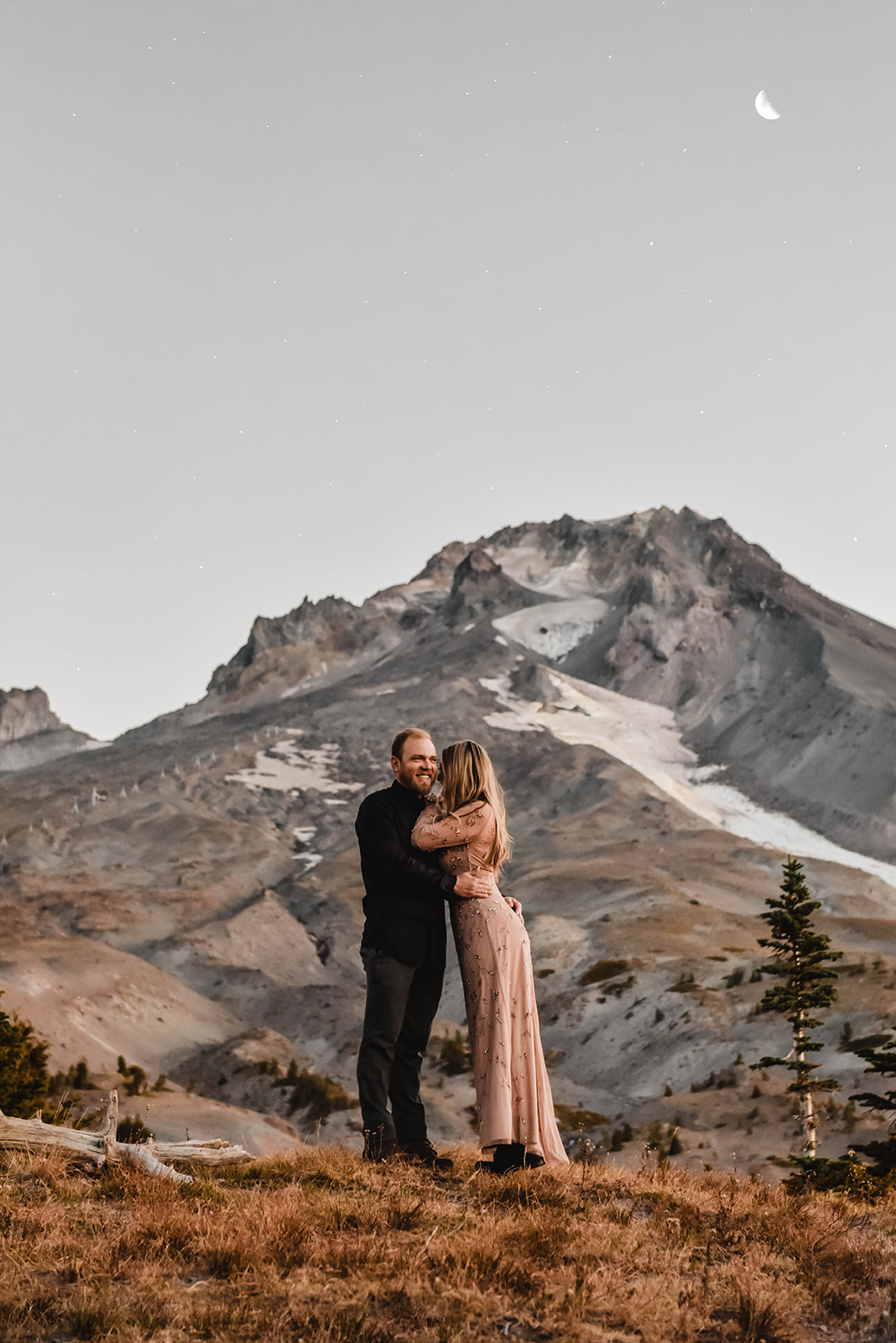 A couple standing in front of Mt. Hood with the moon behind them. 