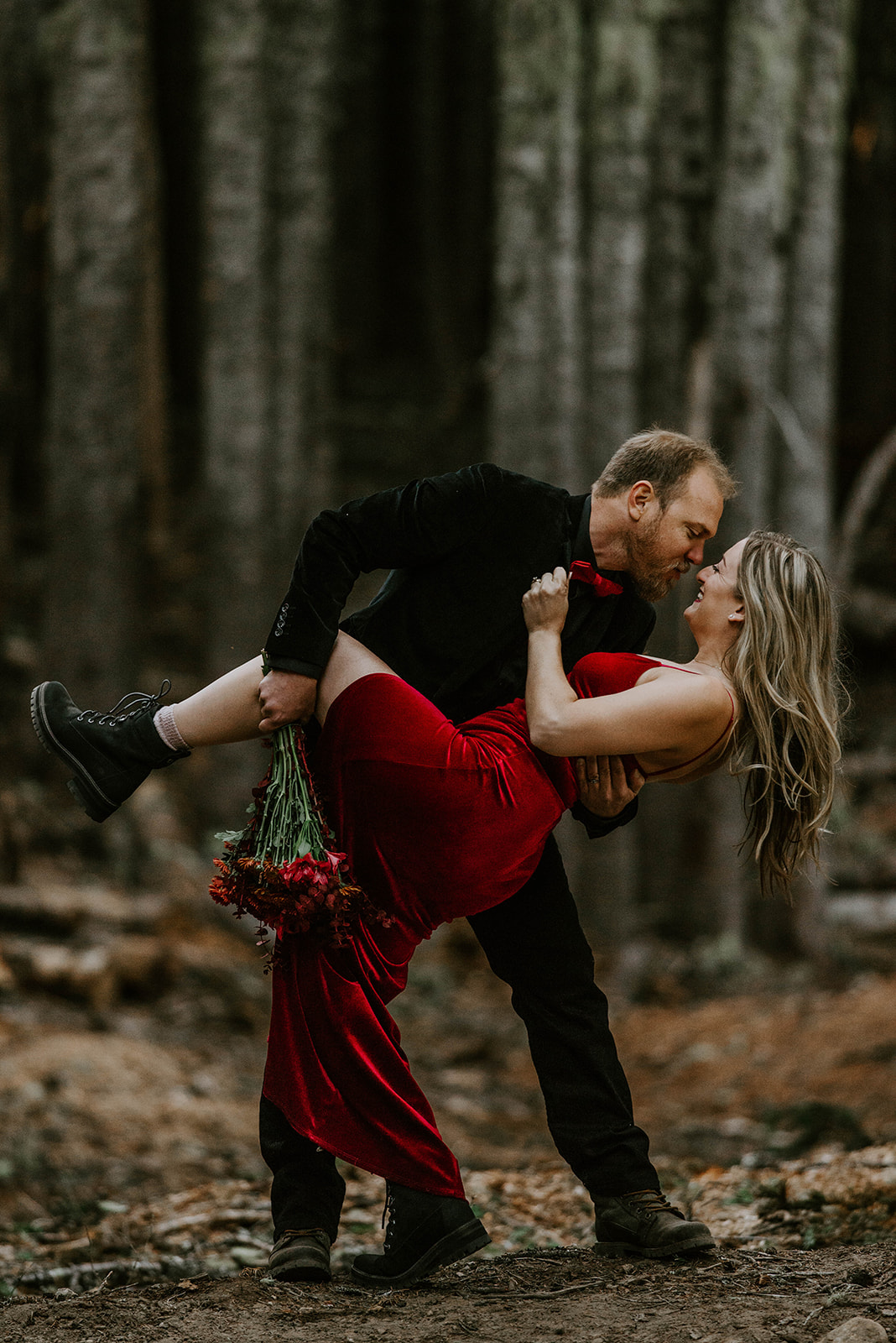 A man dipping his wife in the forest while she wears a red velvet dress for engagement photos