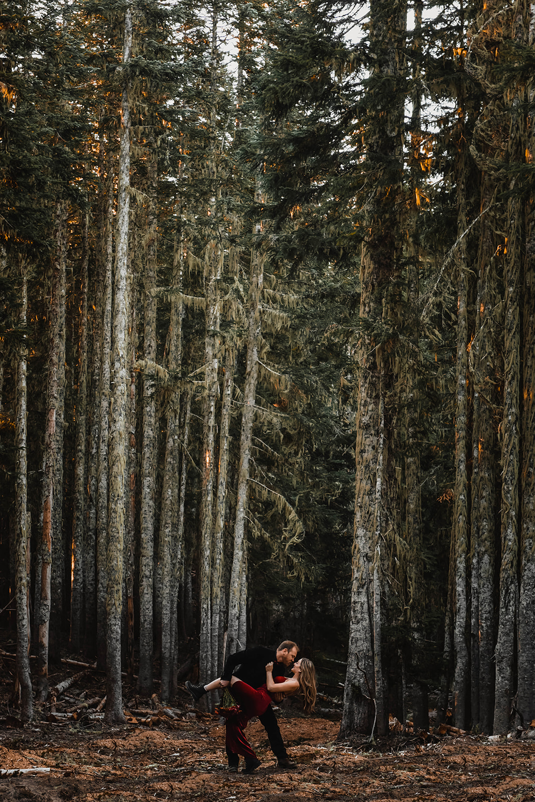 A man dipping his wife in the forest while she wears a red velvet dress for engagement photos