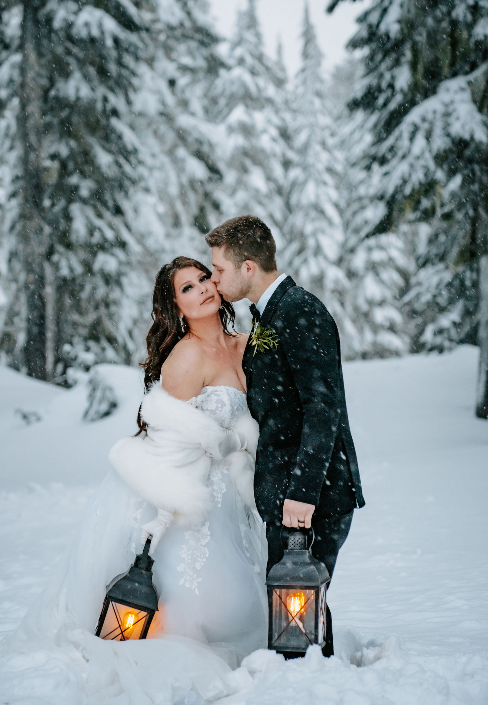 Groom kissing bride's cheek while they hold lanterns in a snowstorm during a winter elopement on Mt. Hood in Oregon.
