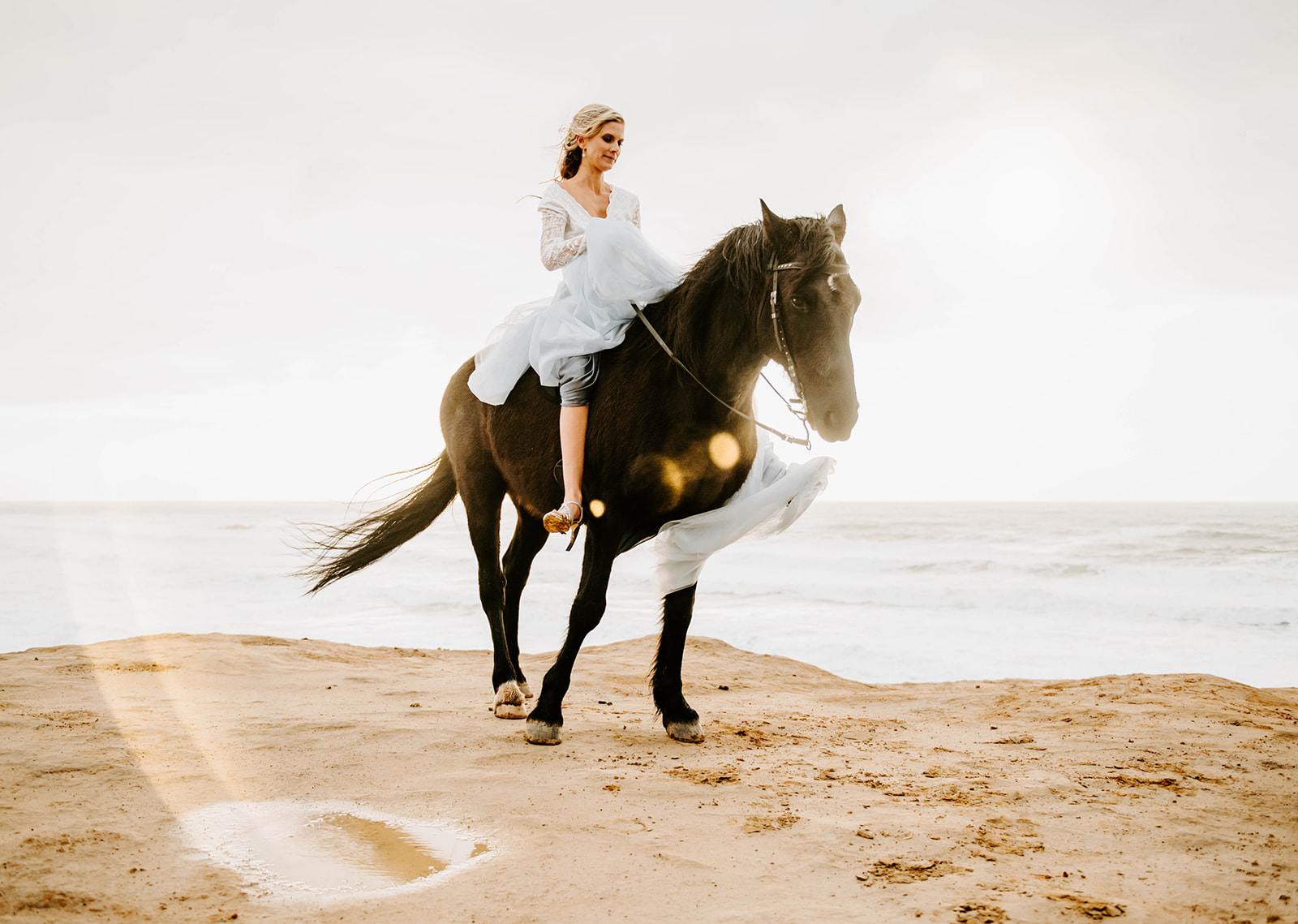 A blonde bride in a blue dress, riding her black horse on the rainy Oregon Coast.