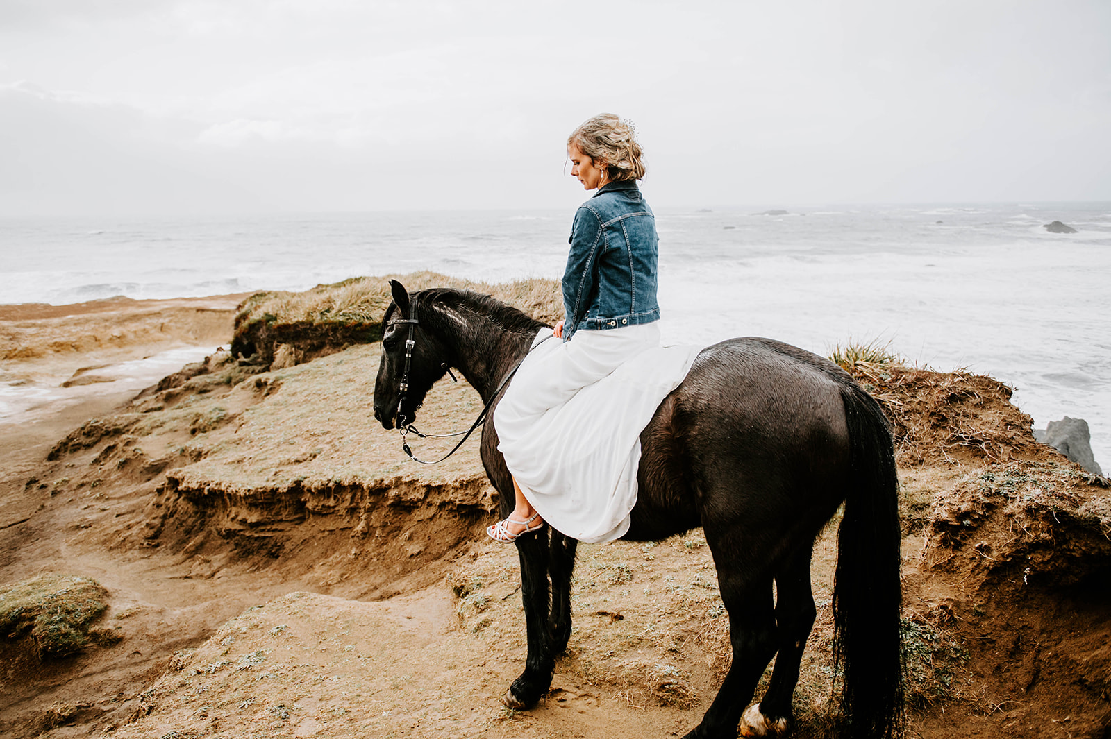 A blonde bride in a jean jacket riding her black horse in her wedding dress on the rainy Oregon Coast.
