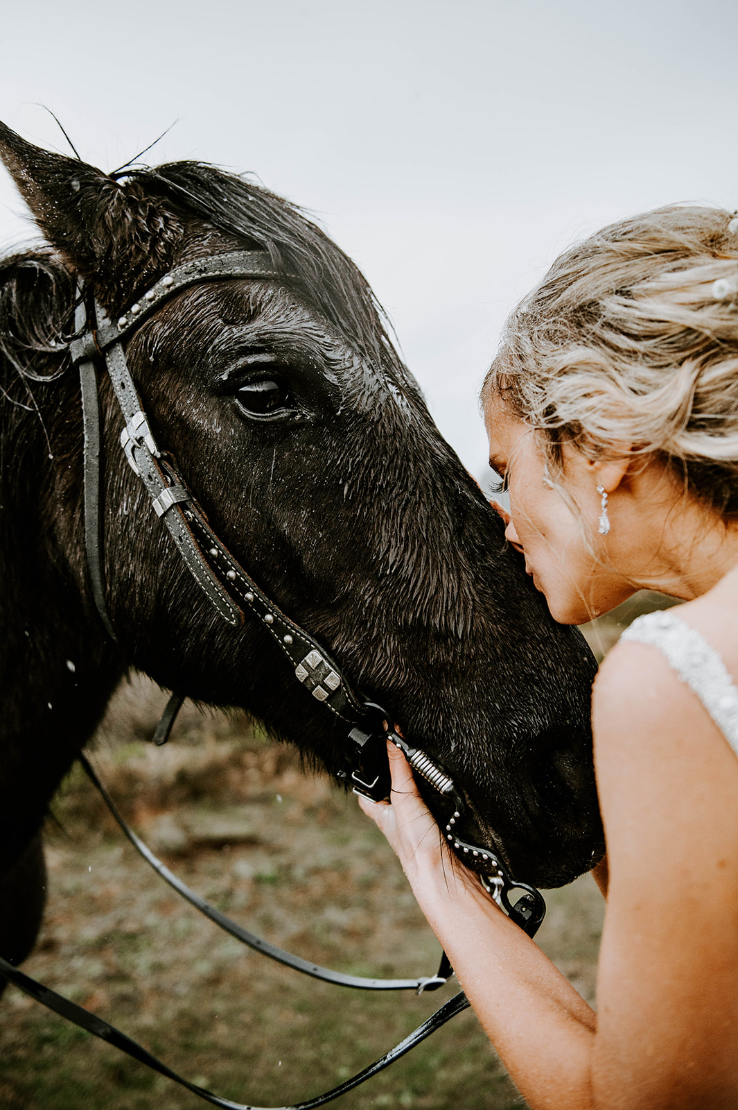 A blonde bride kissing her black horse on the nose while it rains.
