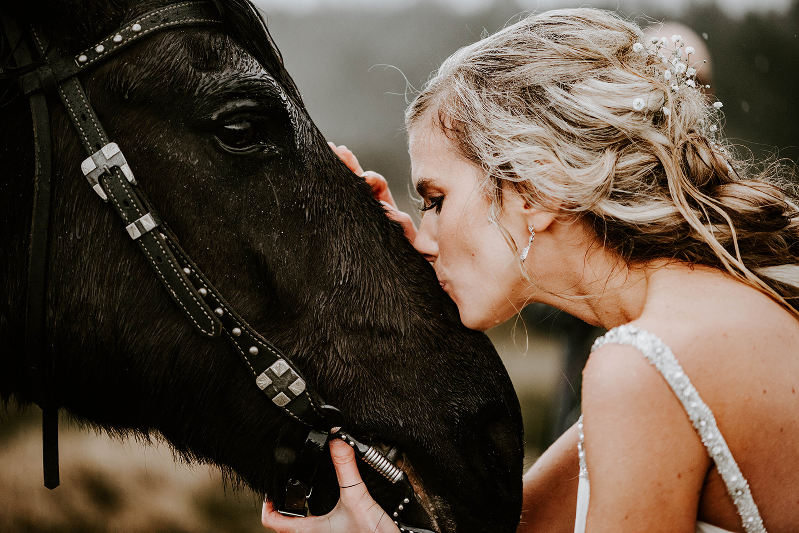A blonde bride kissing her black horse on the nose while it rains.