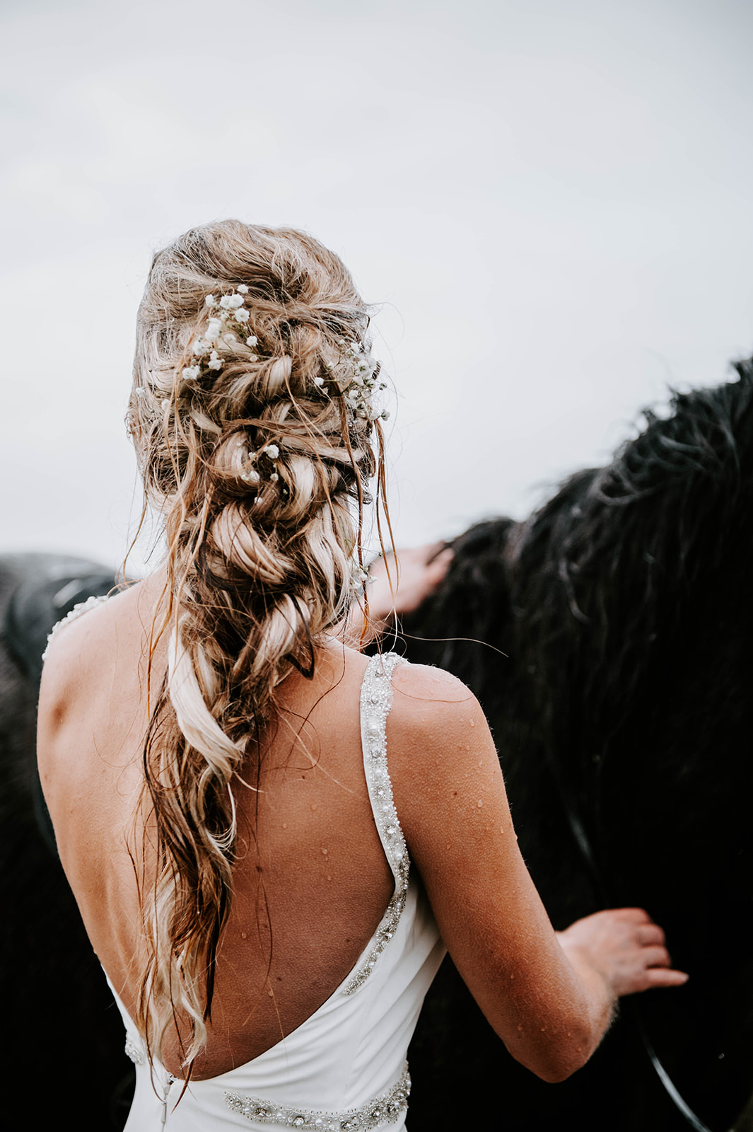 A blonde bride with a messy loose half braid updo while she pets her horse in the pouring rain on the Oregon Coast.