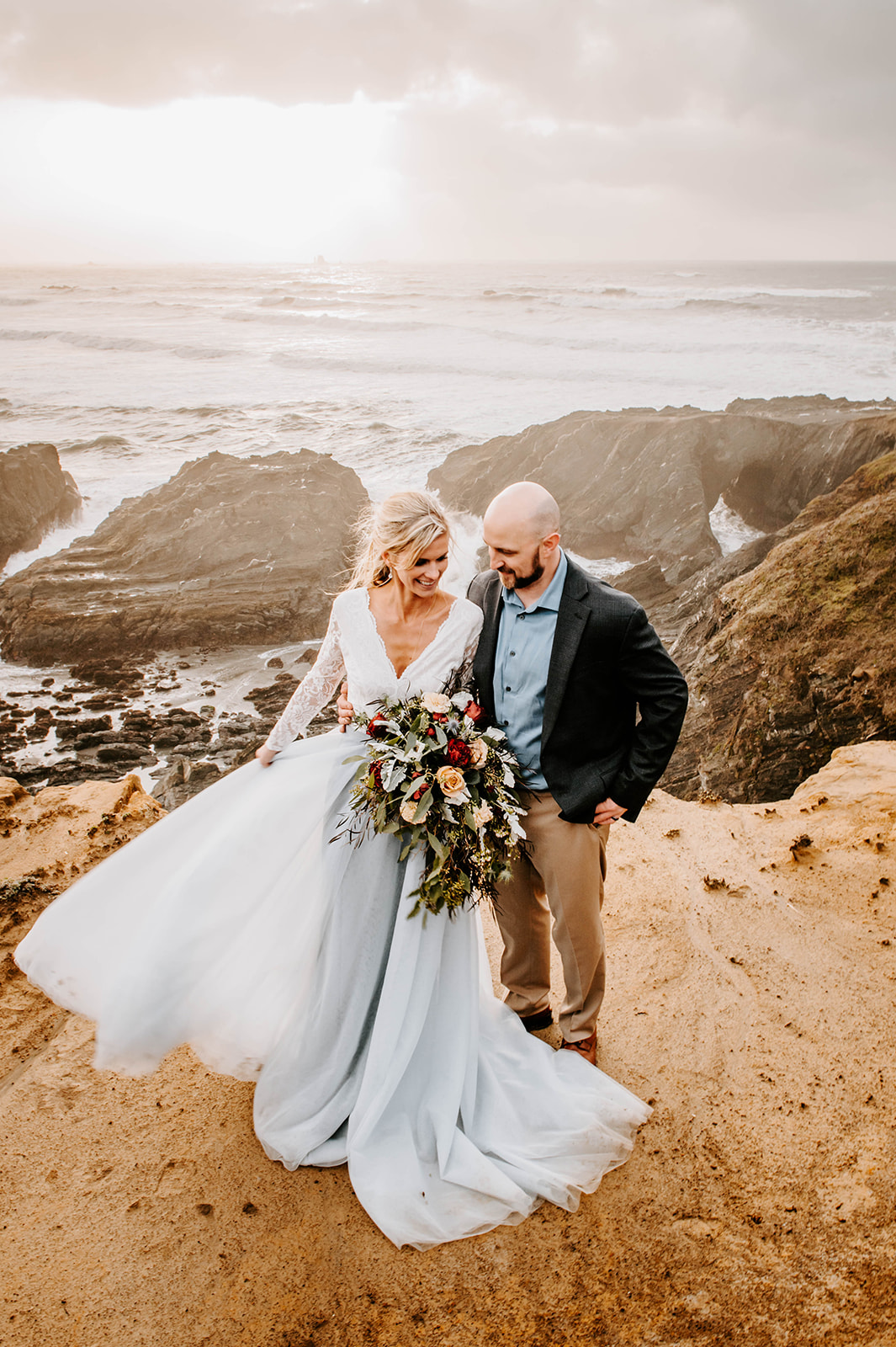 A bride in a blue dress and a groom in khakis, a blue shirt, and a black blazer, overlooking the waves on Otter Point.