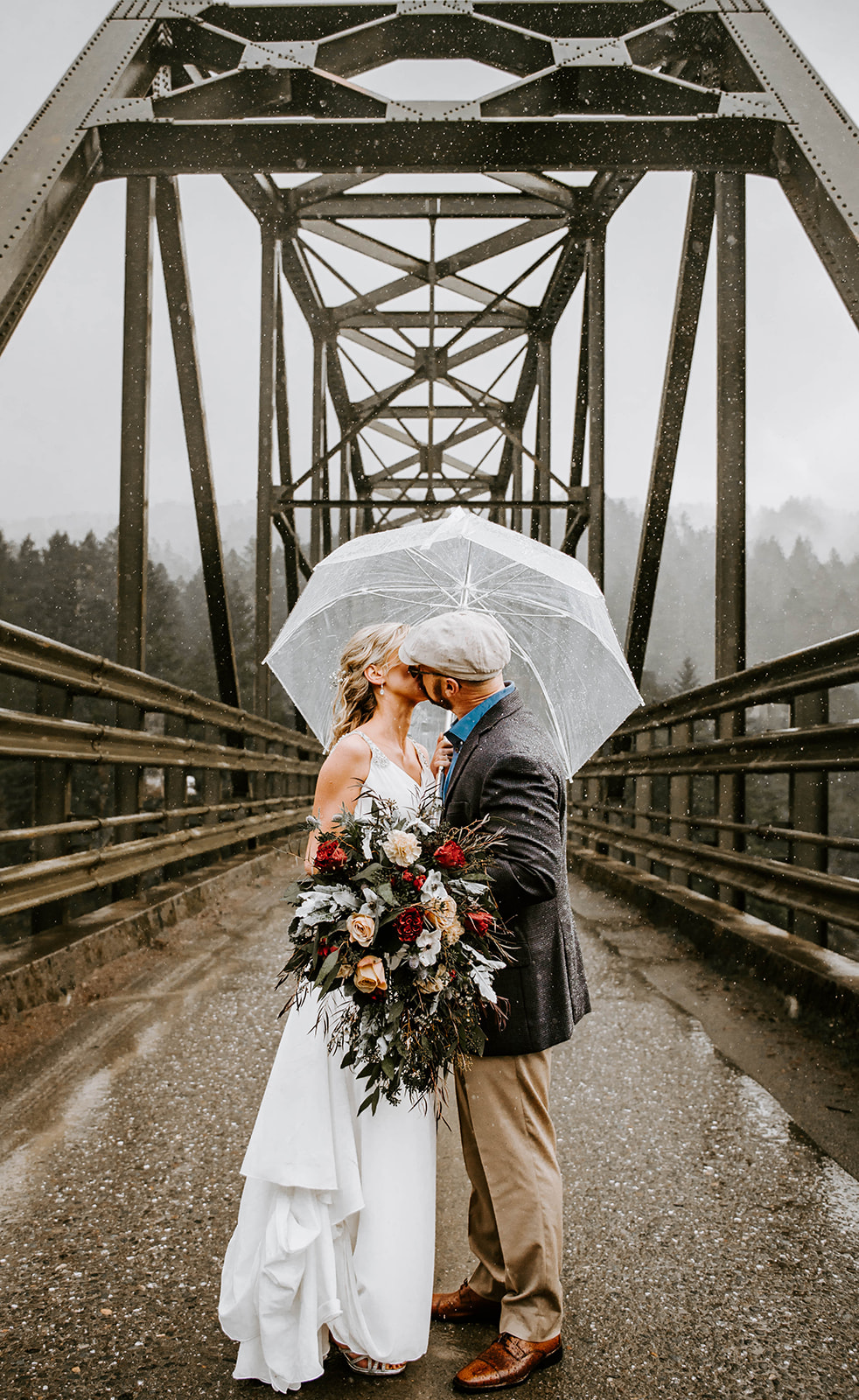 A bride and groom kissing underneath a clear umbrella on a bridge during their winter wedding on the Oregon Coast.
