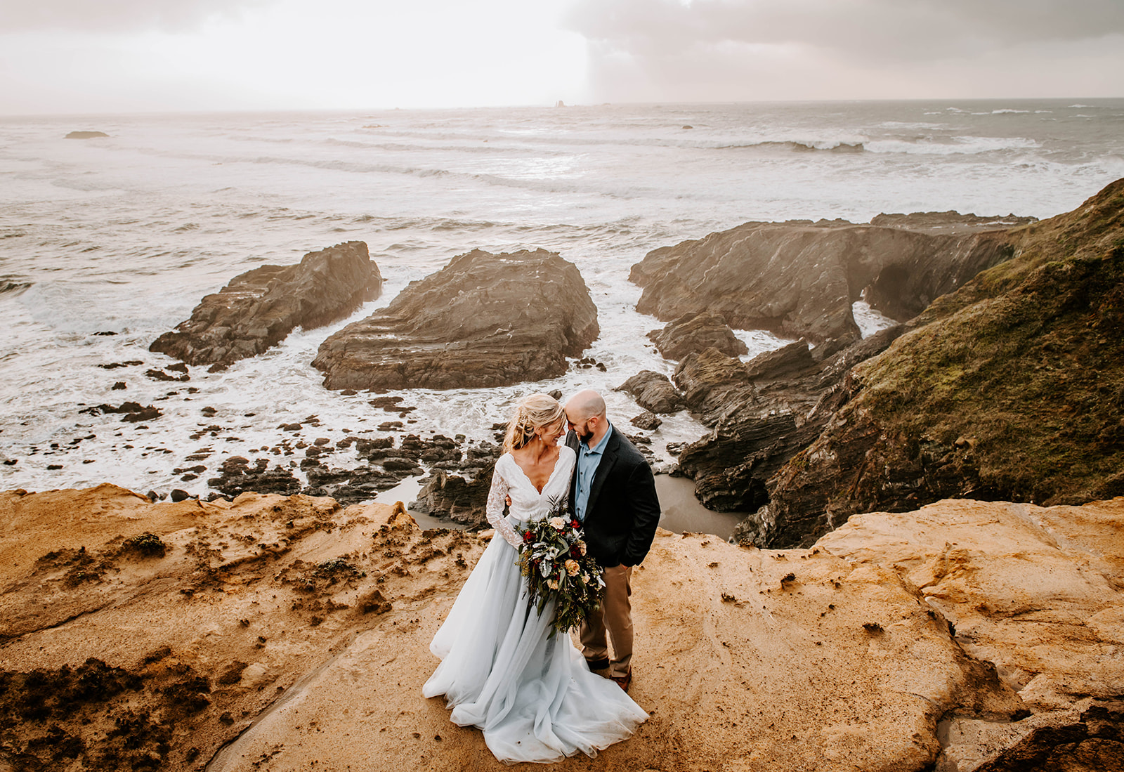 A bride and groom standing forehead to forehead on the cliffs at Otter Point on the Oregon Coast.