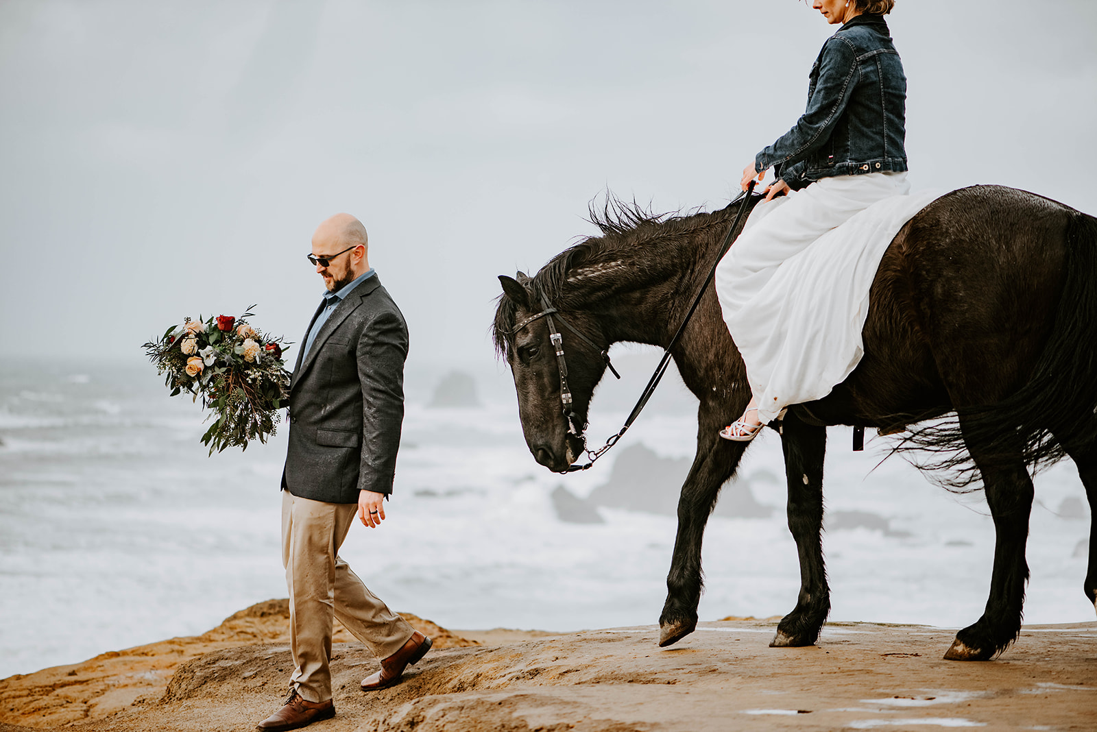 A bride in a jean jacket riding her horse in her wedding dress on the rainy Oregon Coast while groom carries bouquet