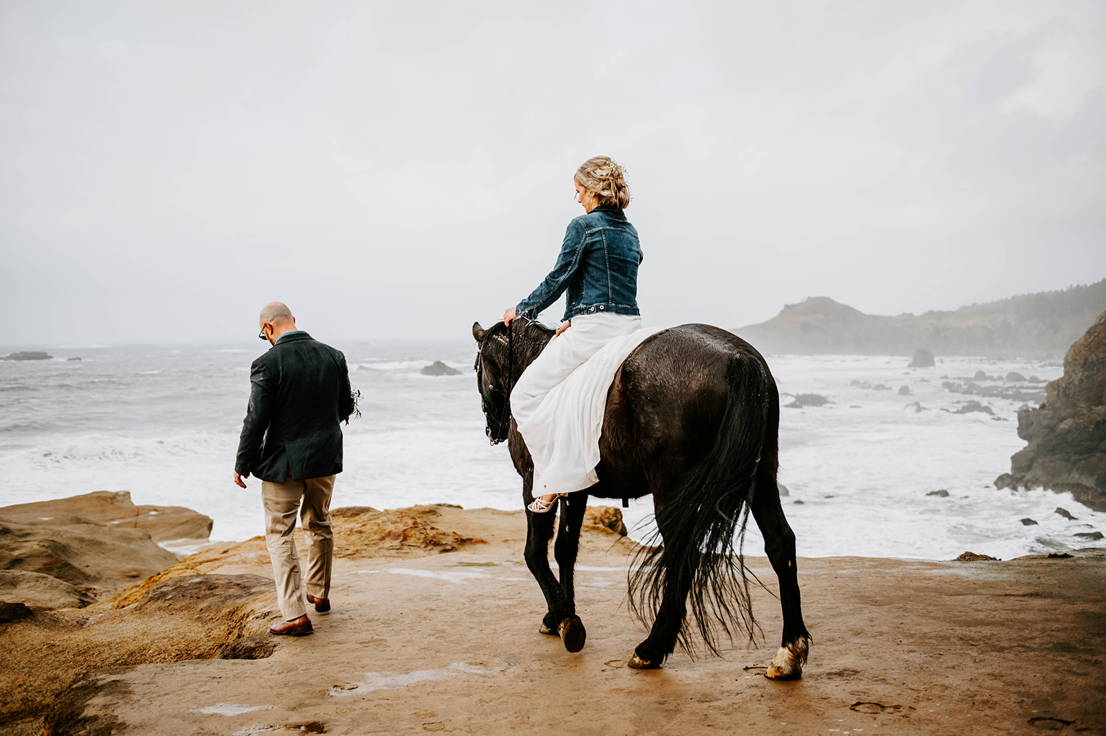 A bride in a jean jacket riding her horse in her wedding dress on the rainy Oregon Coast while groom walks