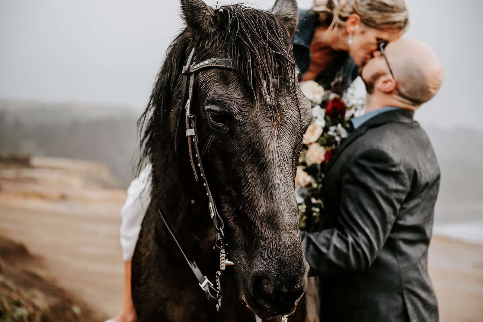 A bride leaning down to kiss the groom while she rides a horse on their wedding day while it rains.