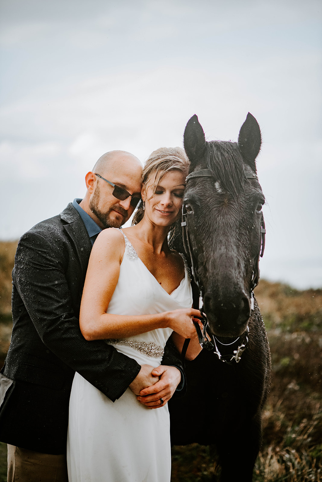 A bride leaning on her black horse in the rain while the groom hugs her from behind.
