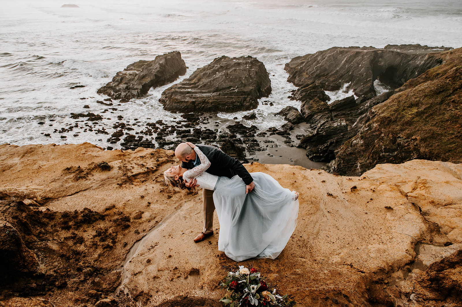 A groom dipping and kissing his bride on the Otter Point Cliffs at the Oregon Coast.