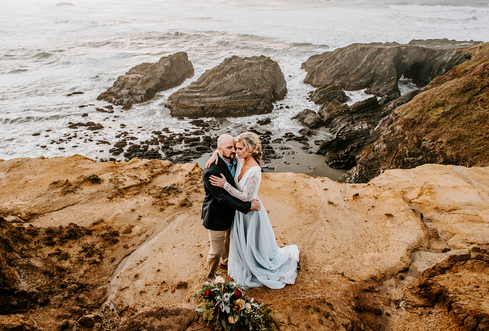 The bride and groom are embracing on the cliffs at Otter Point on the Oregon coast. The bride is wearing a blue dress.