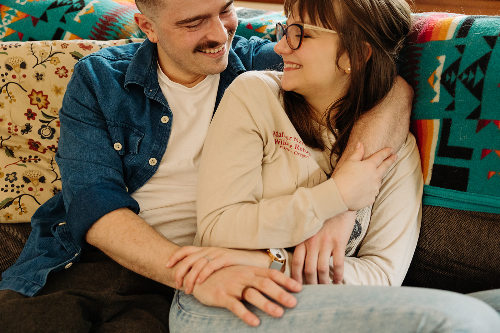 Couple snuggles one another on their couch during their at home engagement photo session