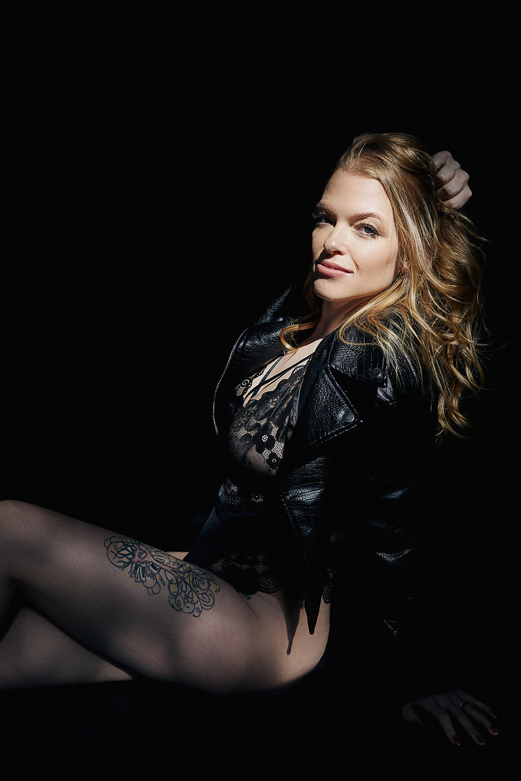 Boudoir photograph of a woman with red hair sitting on the ground in the sun wearing a black leather jacket and lace