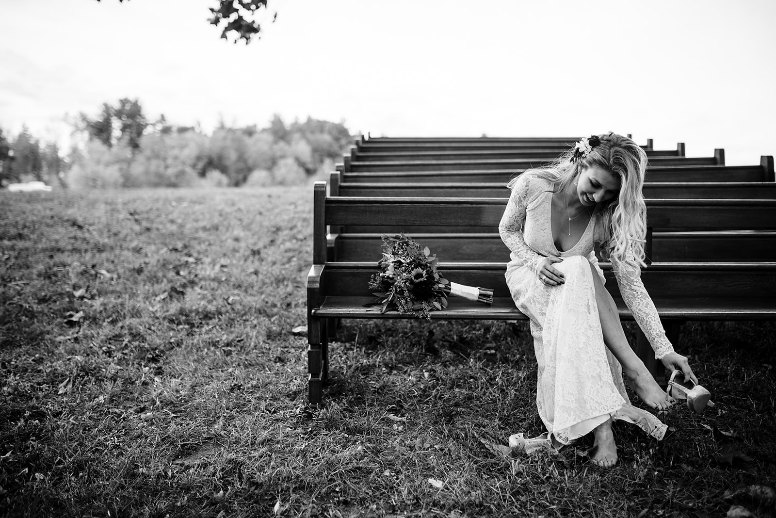 Bride sits on wooden pew to take her shoes off with her bouquet sitting next to her.