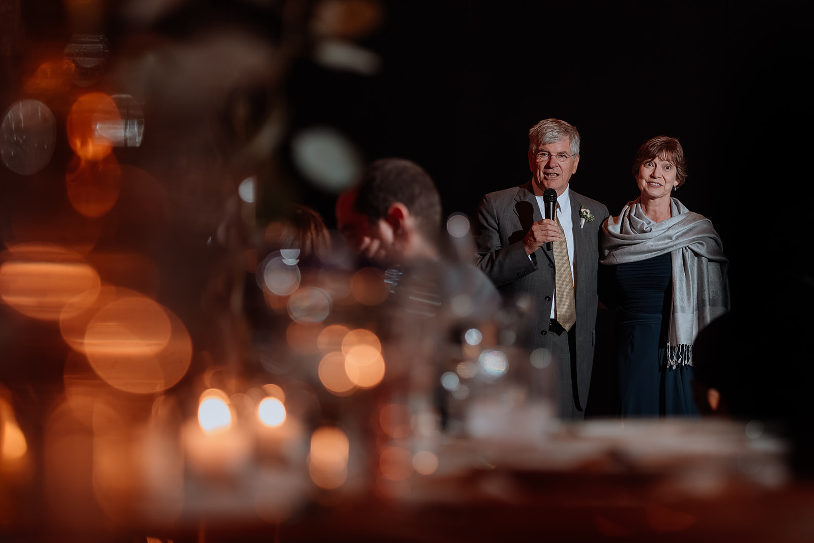 Bride's Parents are framed with the orange bokeh from shallow depth of view while giving a toast at reception.