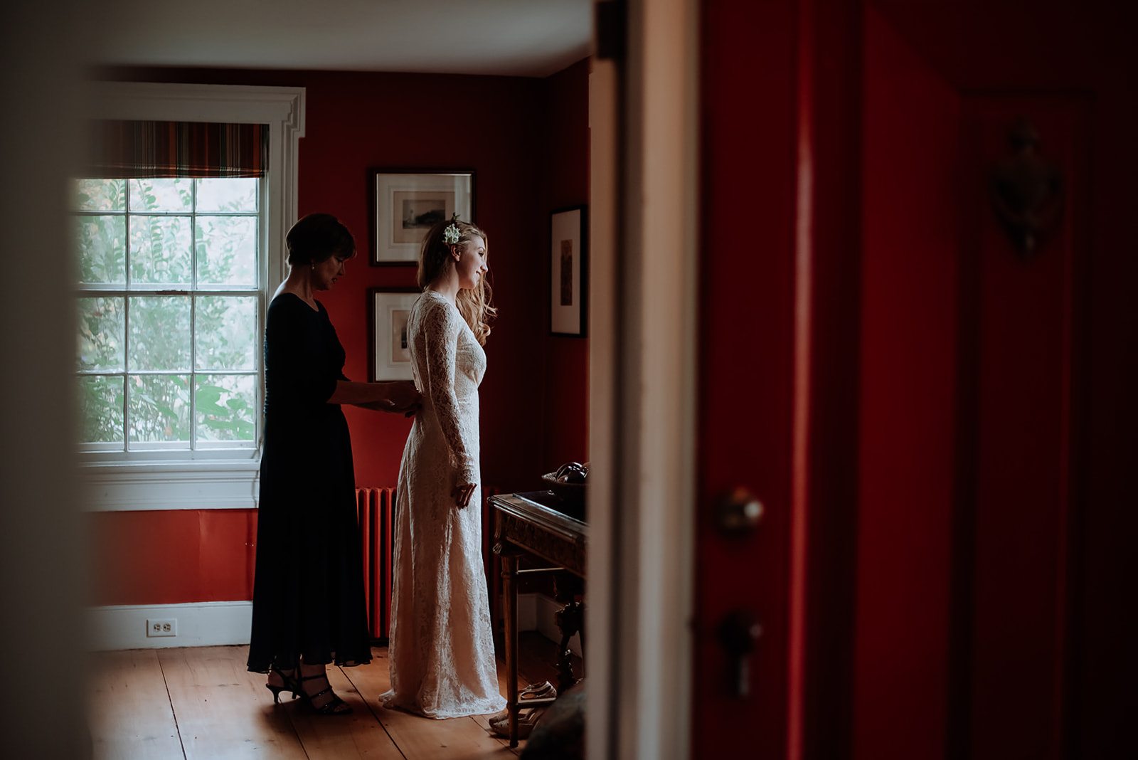 Brides mother sips up her daughters vintage lace thrifted dress while standing in a red living room of the farmhouse. 