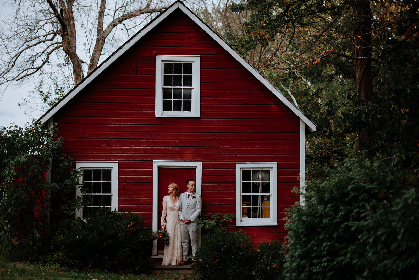 Couple poses in front of red barn on a cloudy day wedding in Hudson Valley New York.