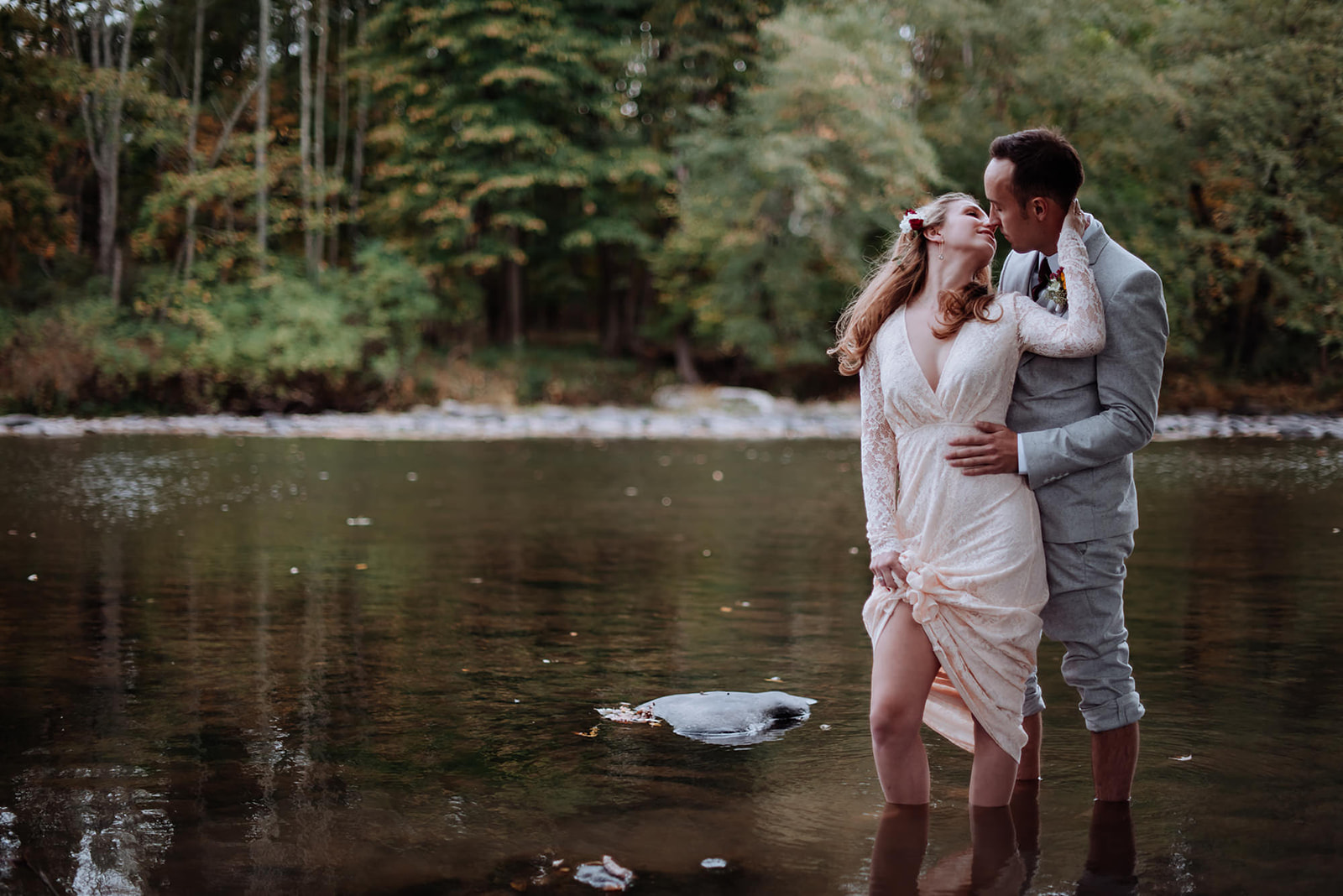 Couple romantically stand in shallow water while bride reaches back to caress her husbands face.