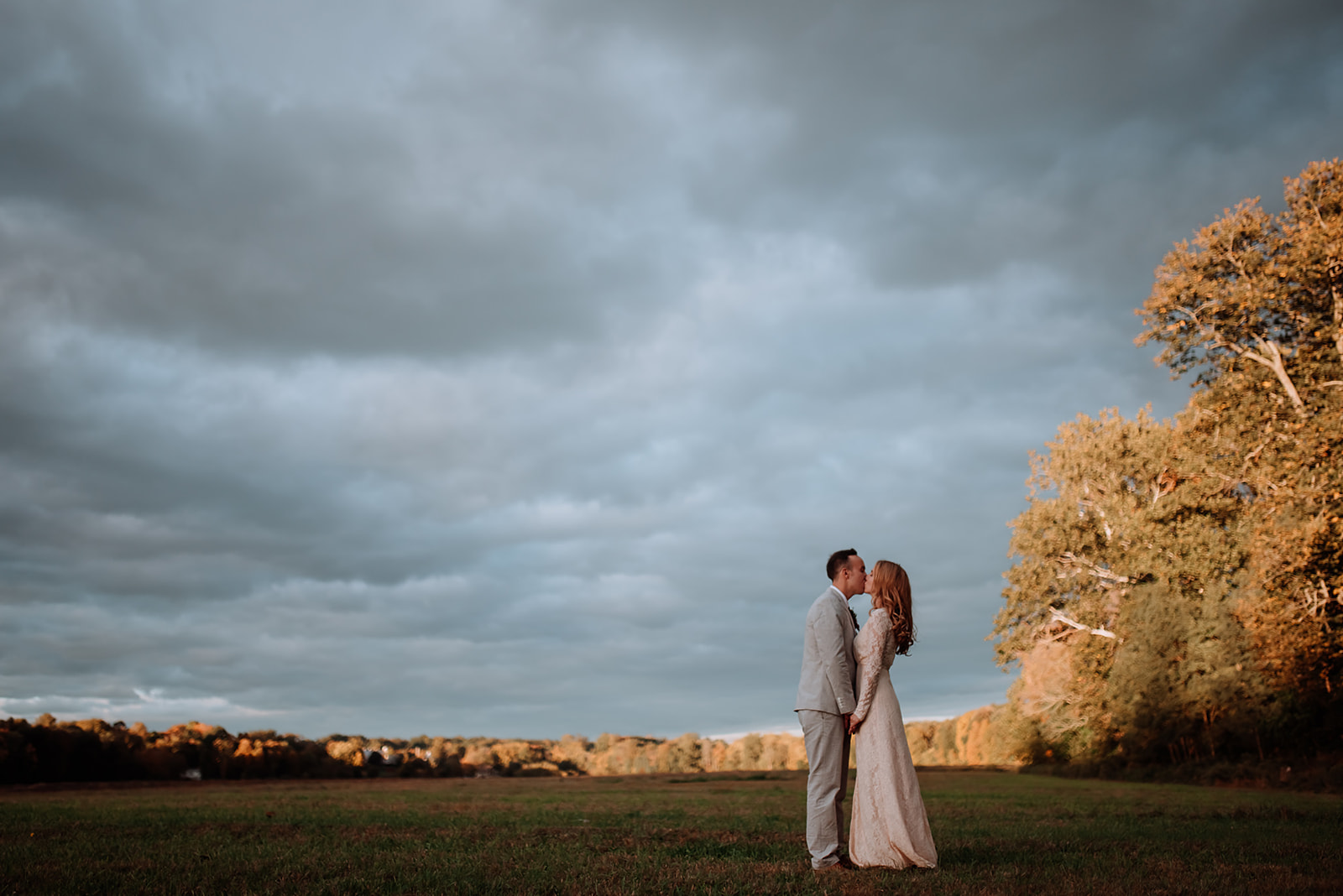 Couple shares a sweet kiss during golden hour on a cloudy day standing in a farm field.