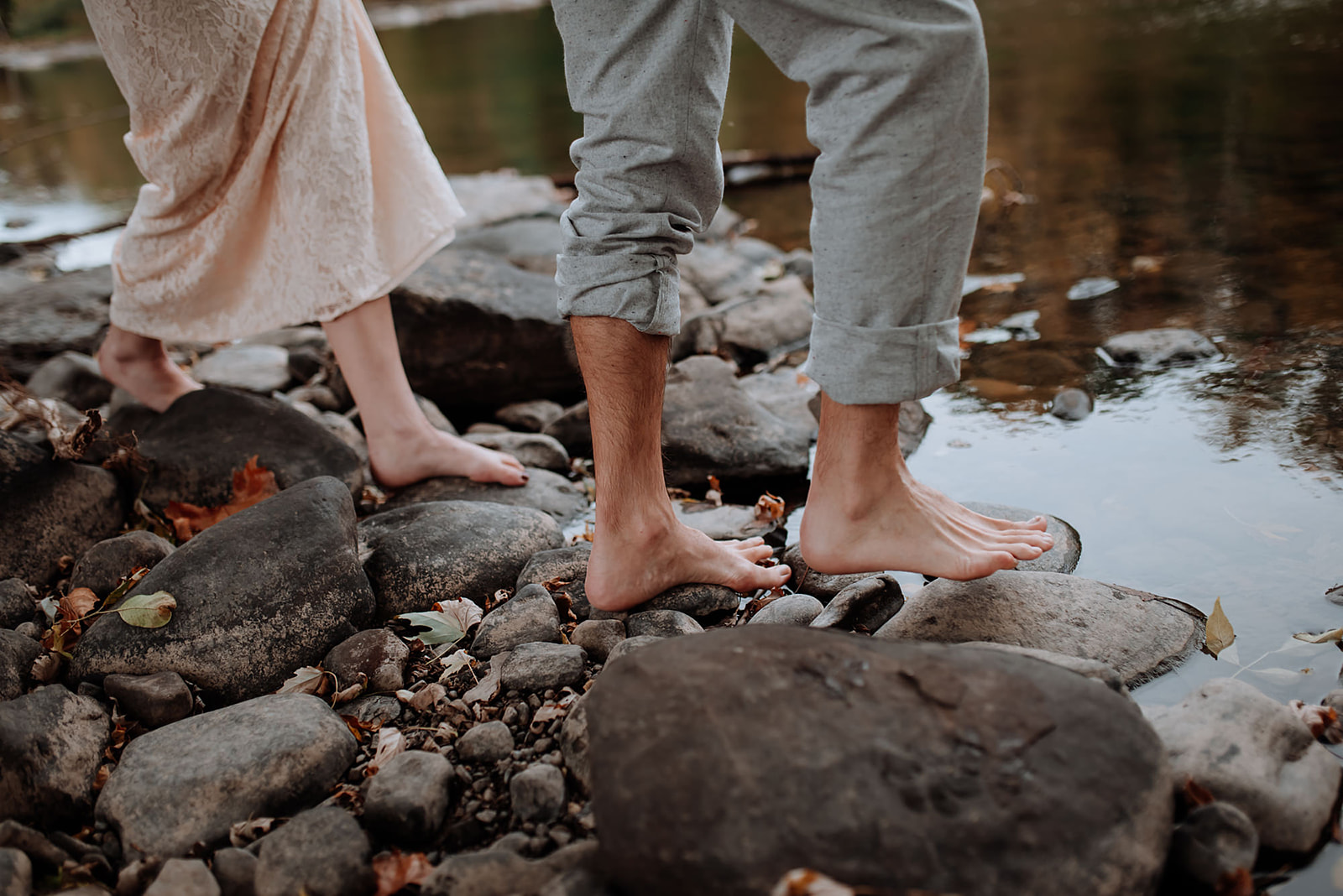 Couples bare feet walk along the rocky rivers edge with reflections of the trees int he water.