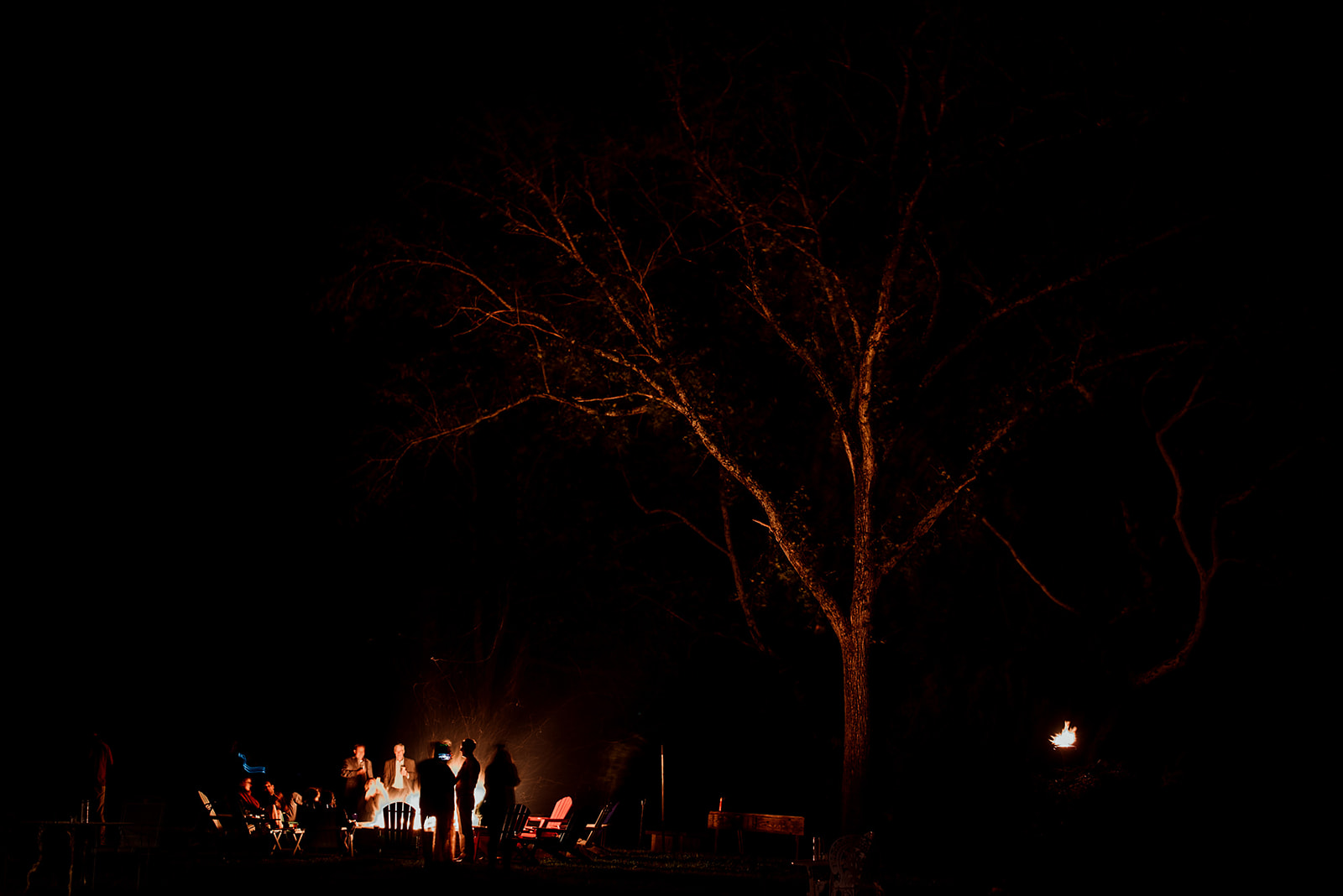 Far away view of after party bon fire with guests sitting in Adirondack chairs roasting smores.