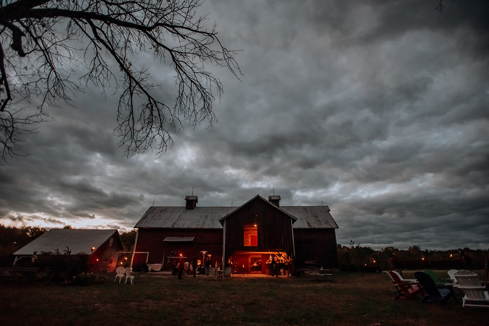 Moody, blue hour, cloudy sky contrasts red barn at dusk with warm orange bistro lights draped inside barn.