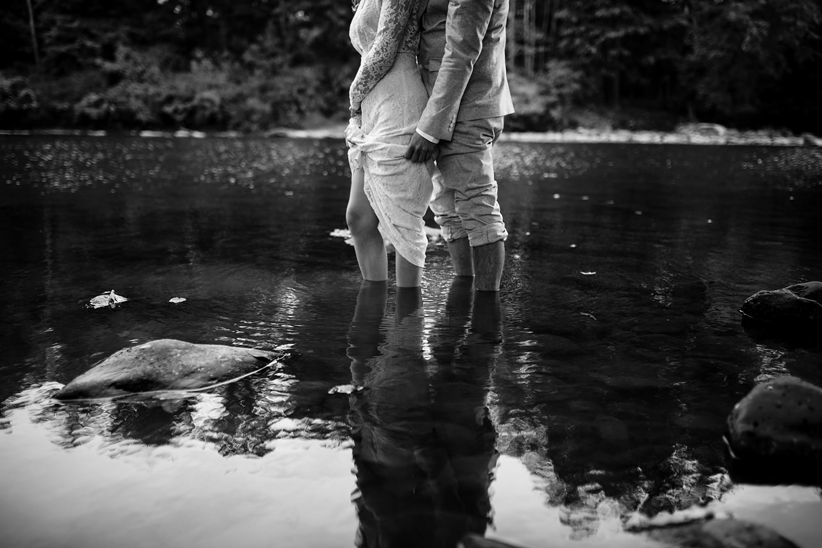 Young hipster couple stands in shallow river while groom holds her dress out of the water at Crested Hen Farms wedding.