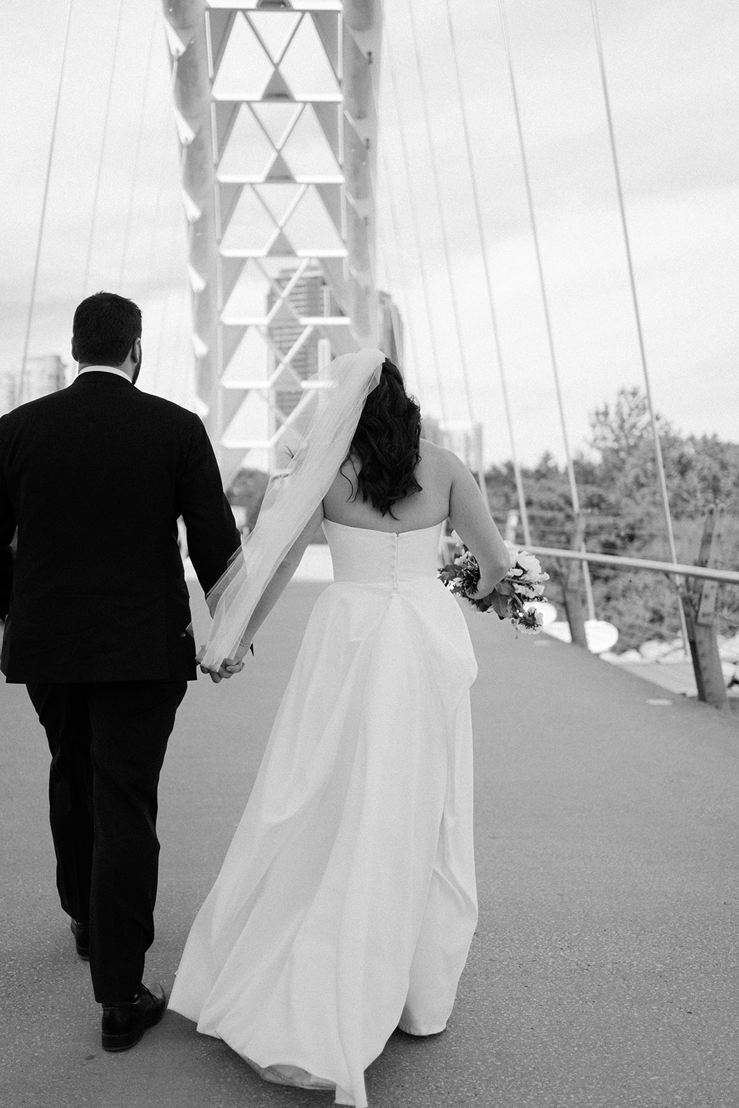 Black and white image of a married couple holding hands walking on a bridge.