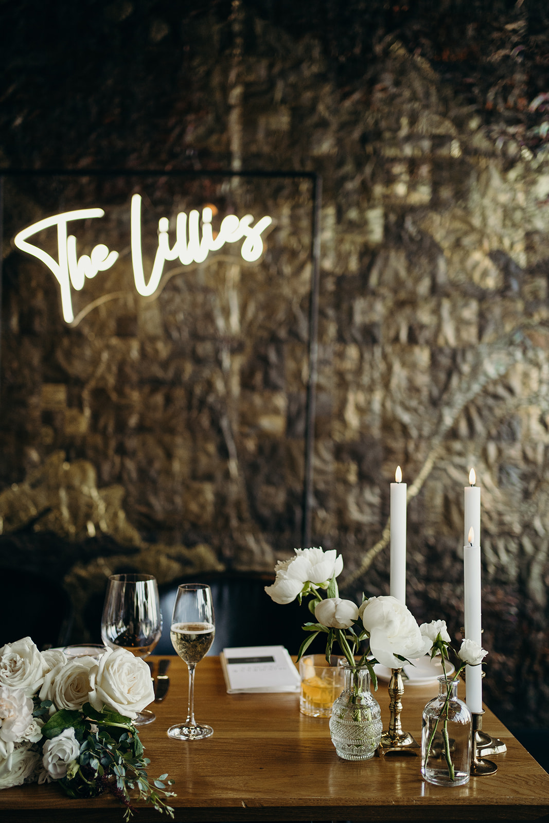 Candles and flowers on the table with a lit up sign hung on a on brick wall.