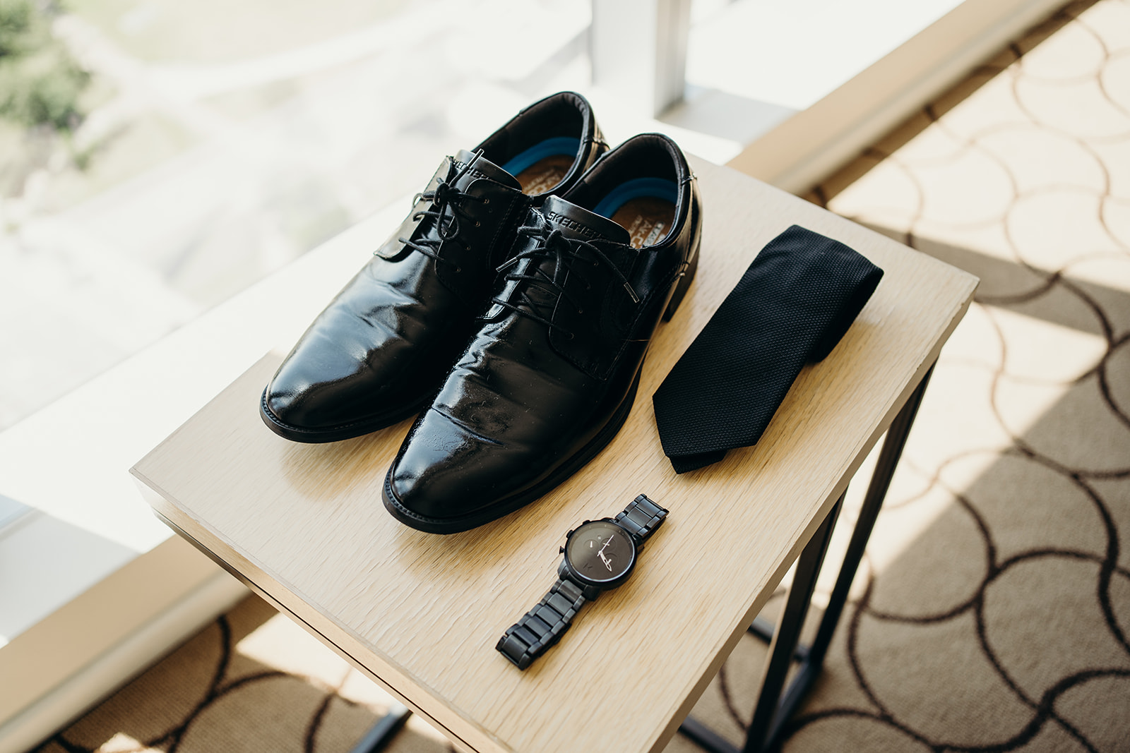 A pair of shoes, a watch and a tie rolled onto a beige square table.