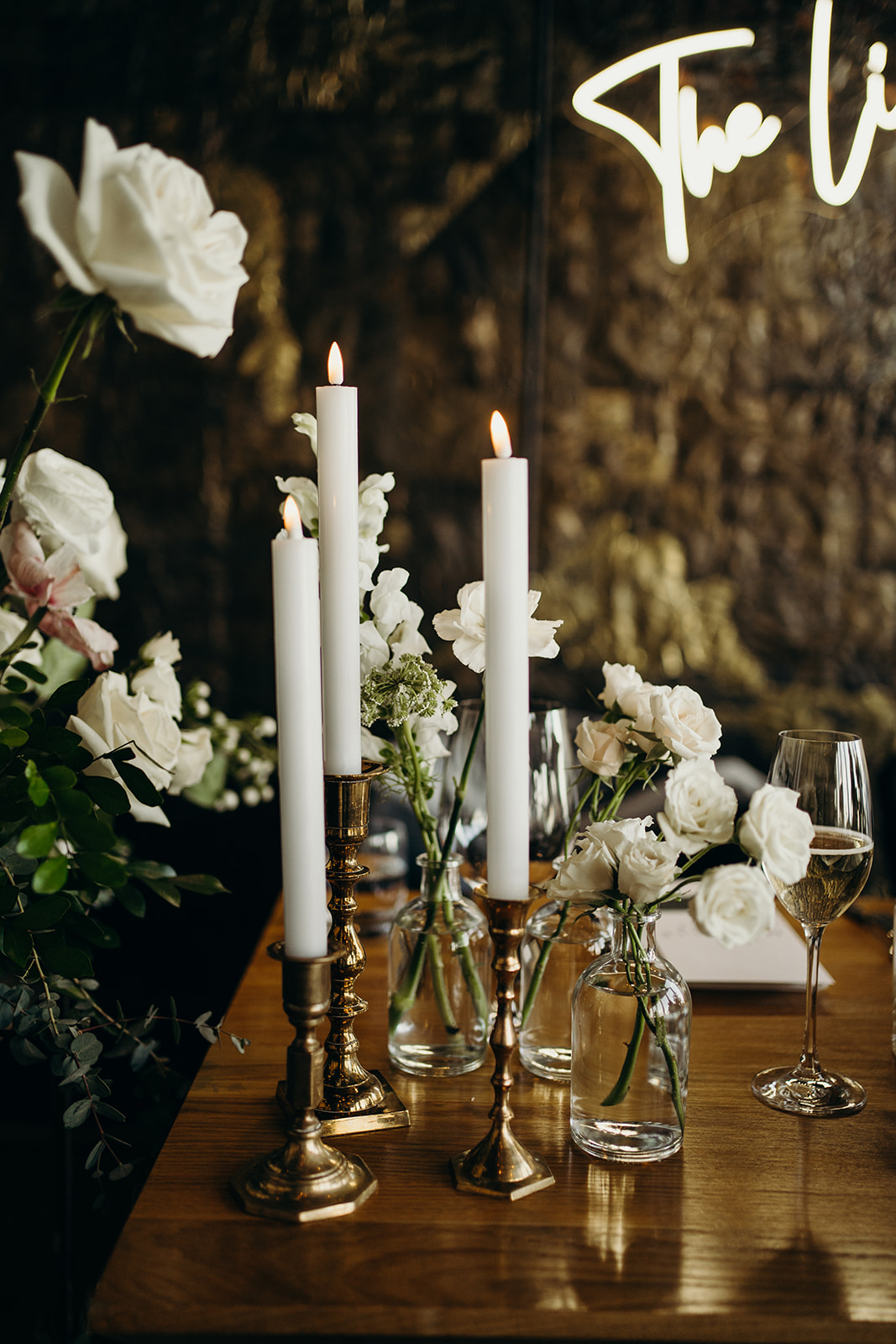 Three lit candles on a table beside flowers in a vase with wine glasses.