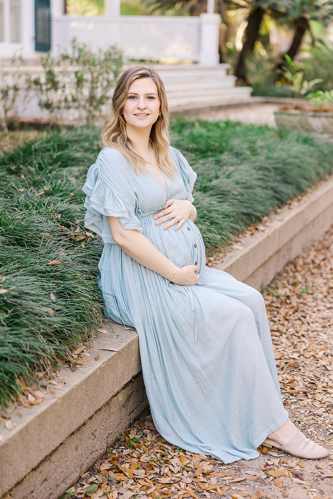 Portrait of woman in blue dress seated for maternity photos with greenery