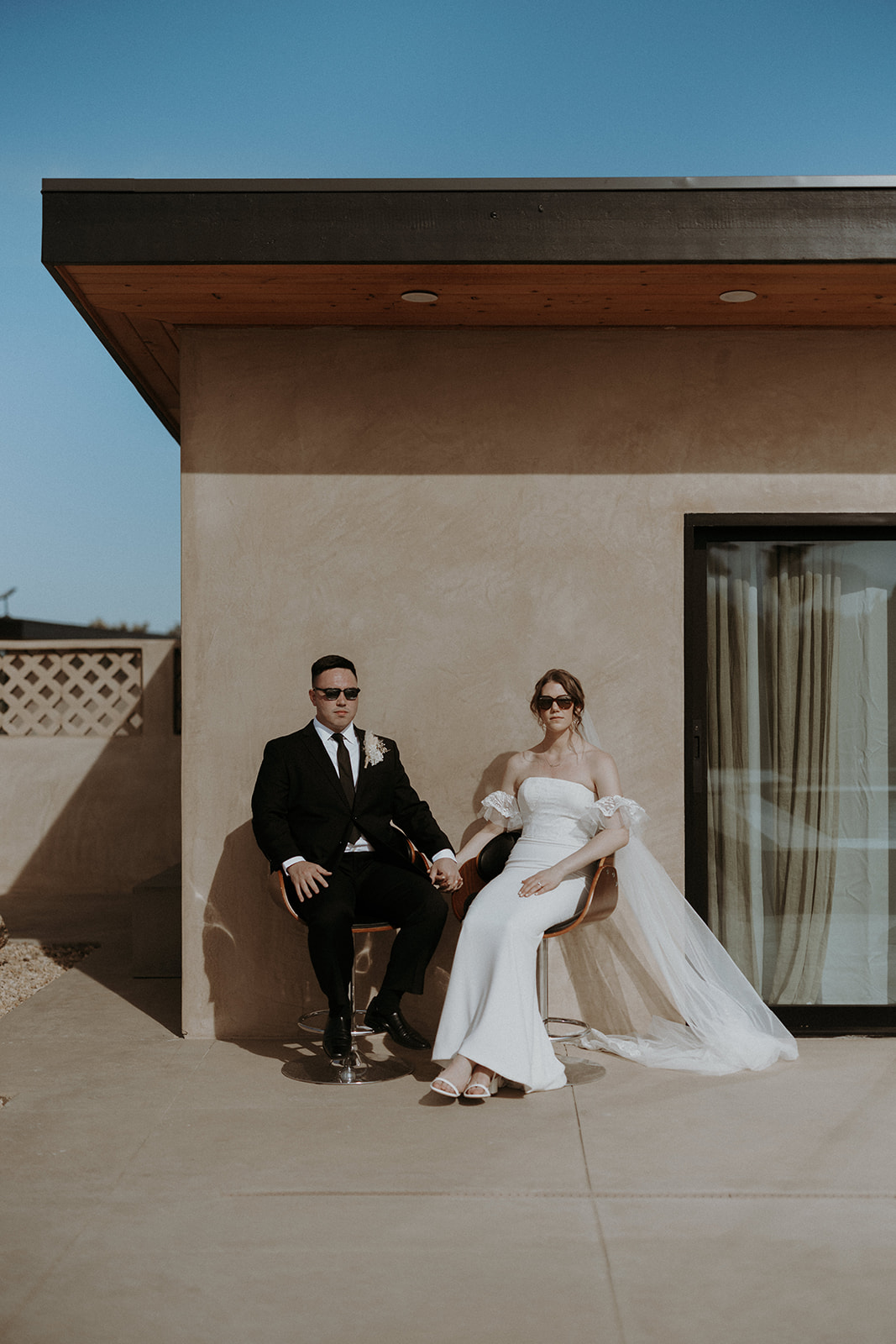 A wedding couple outside a modern boho airbnb sitting on chairs and wearing sunglasses.