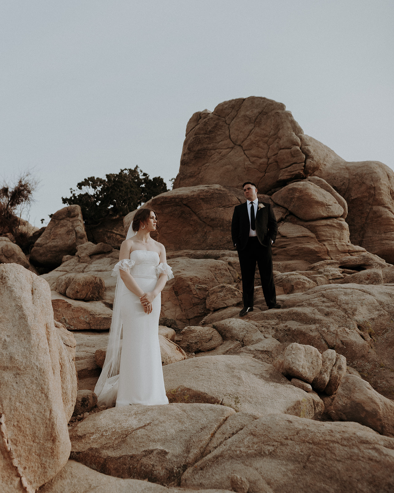 A wedding couple in Joshua Tree National Park standing apart and staggered on a rock formation.