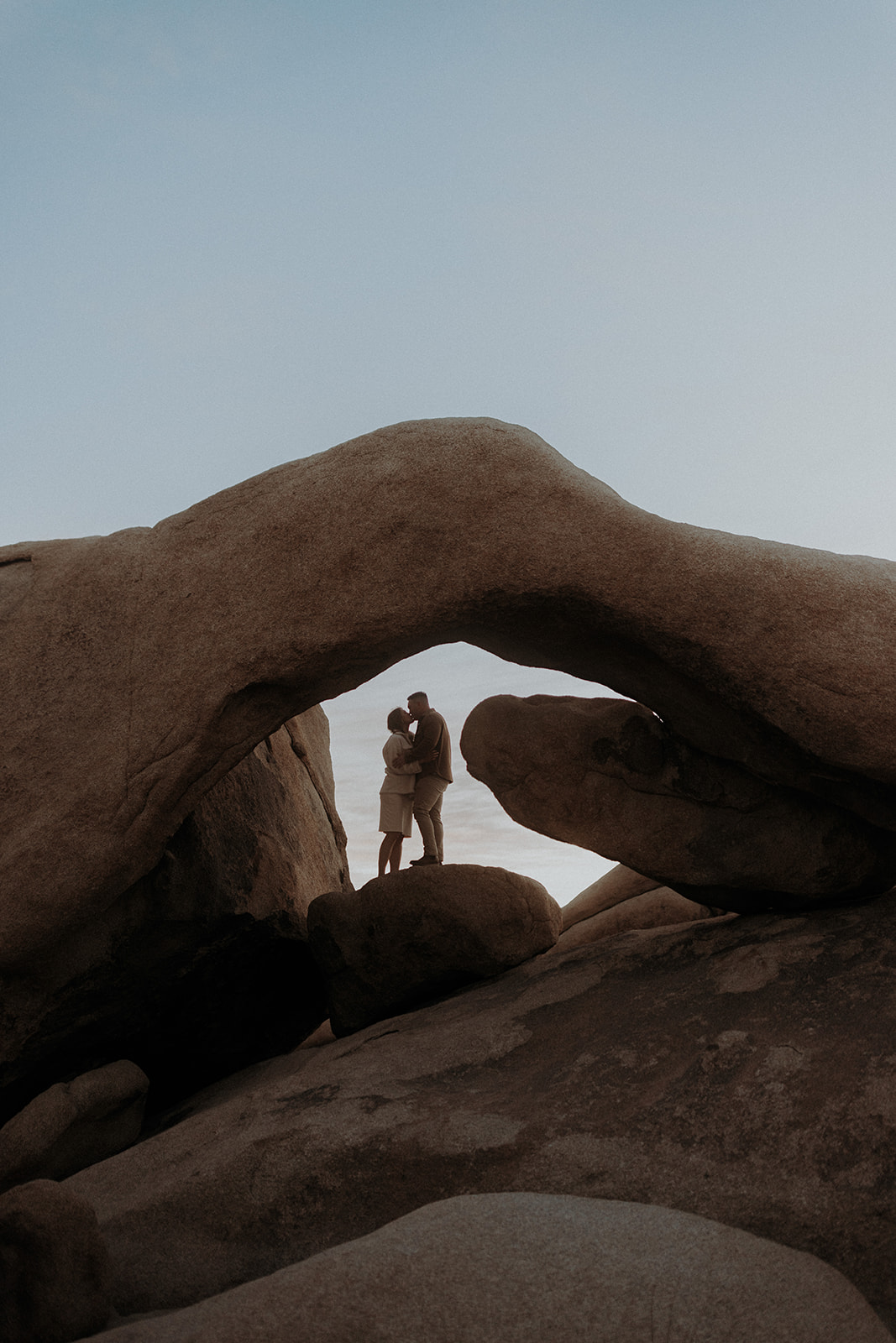 An eloping couple standing under Arch Rock in Joshua Tree National Park at sunrise and kissing.