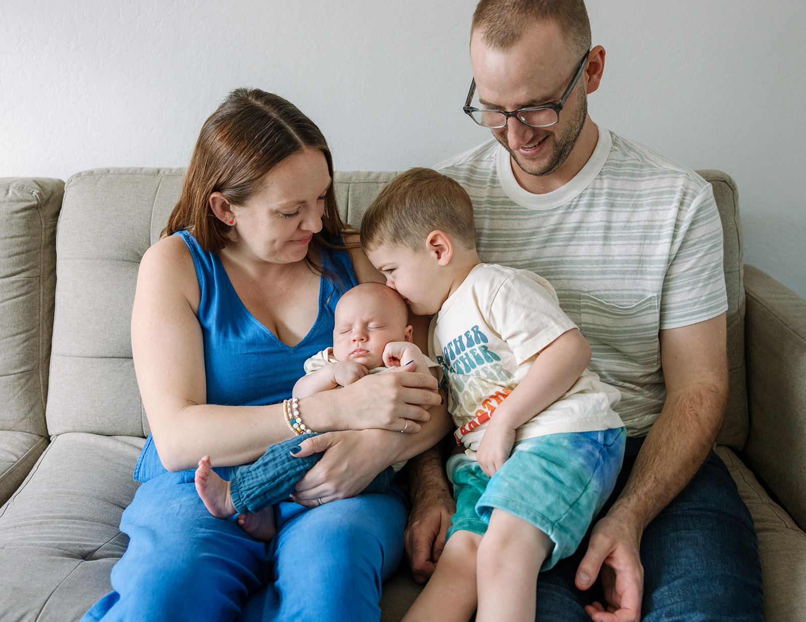 photo of mom and dad sitting on gray couch in living room with toddler son kissing the top of newborn brother's head