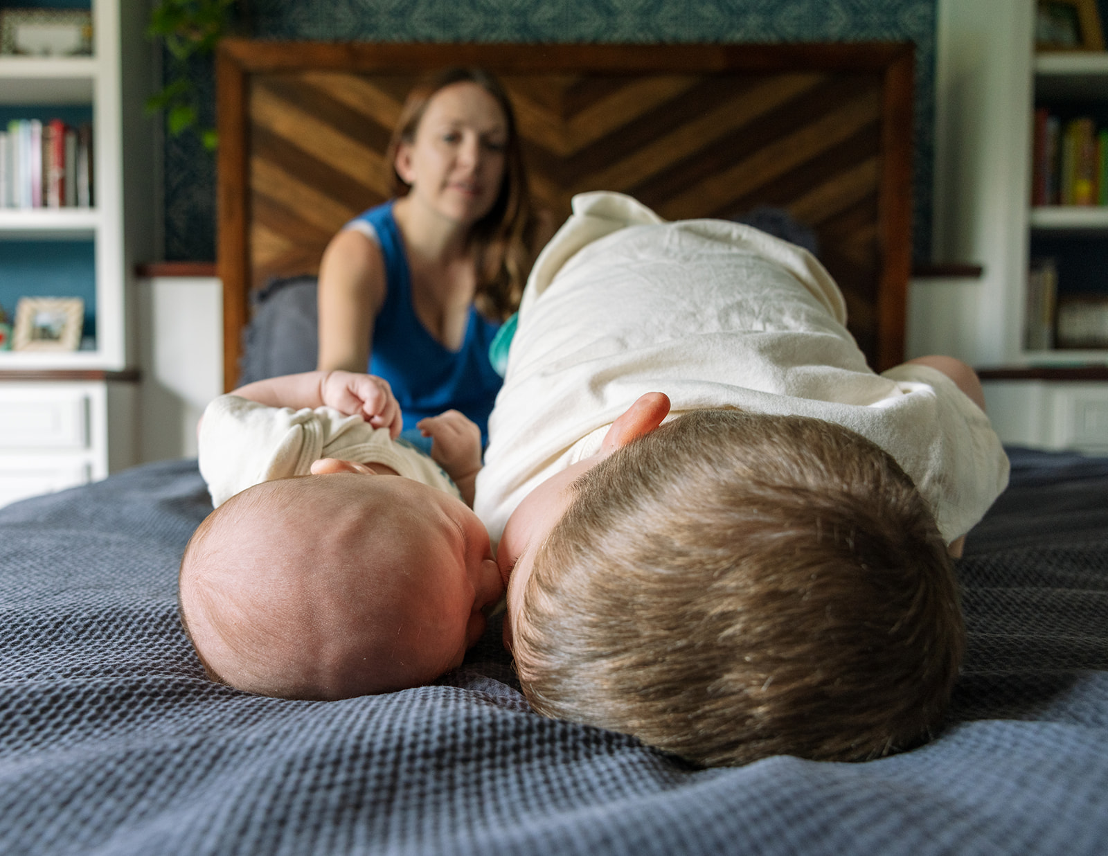 photo of the toddler boy and newborn brother in child's pose on parents' bed while mom looks on in the background