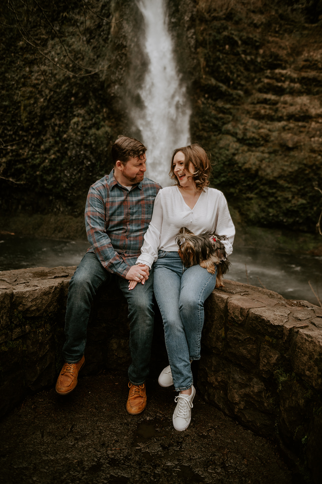 A couple and their dog sitting on a rock wall in front of a portland waterfall for engagement photos